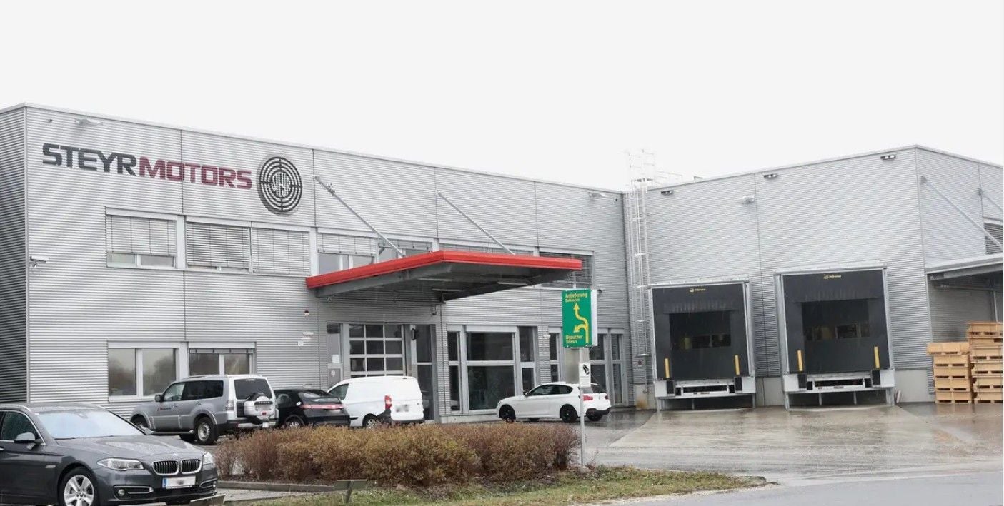 Industriegebäude von Steyr Motors mit geparkten Autos und Ladebuchten bei grauem Himmel.