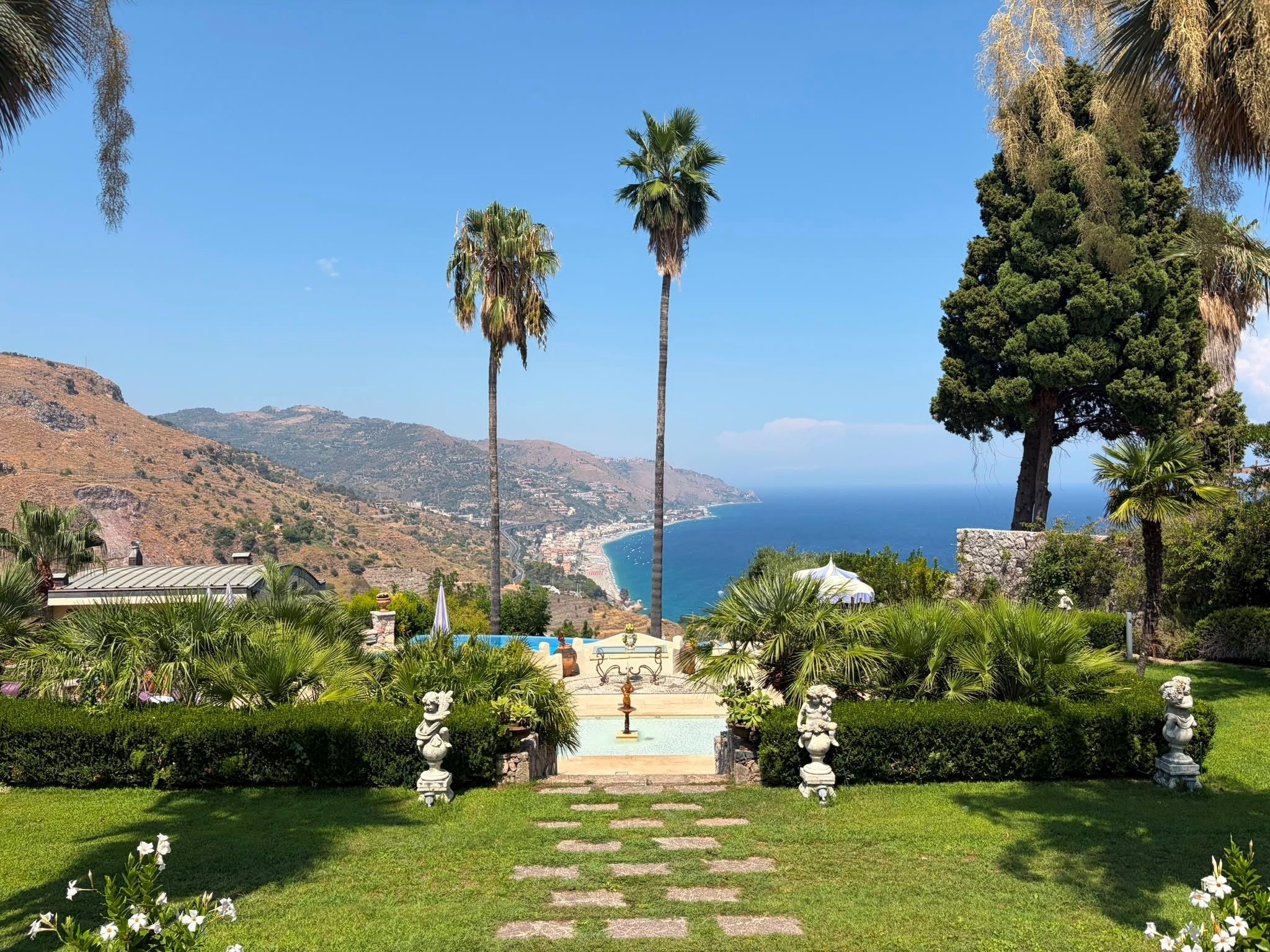 Garten mit Palmen und Statuen, Blick auf Küste und Meer in Sizilien bei klarem Himmel.