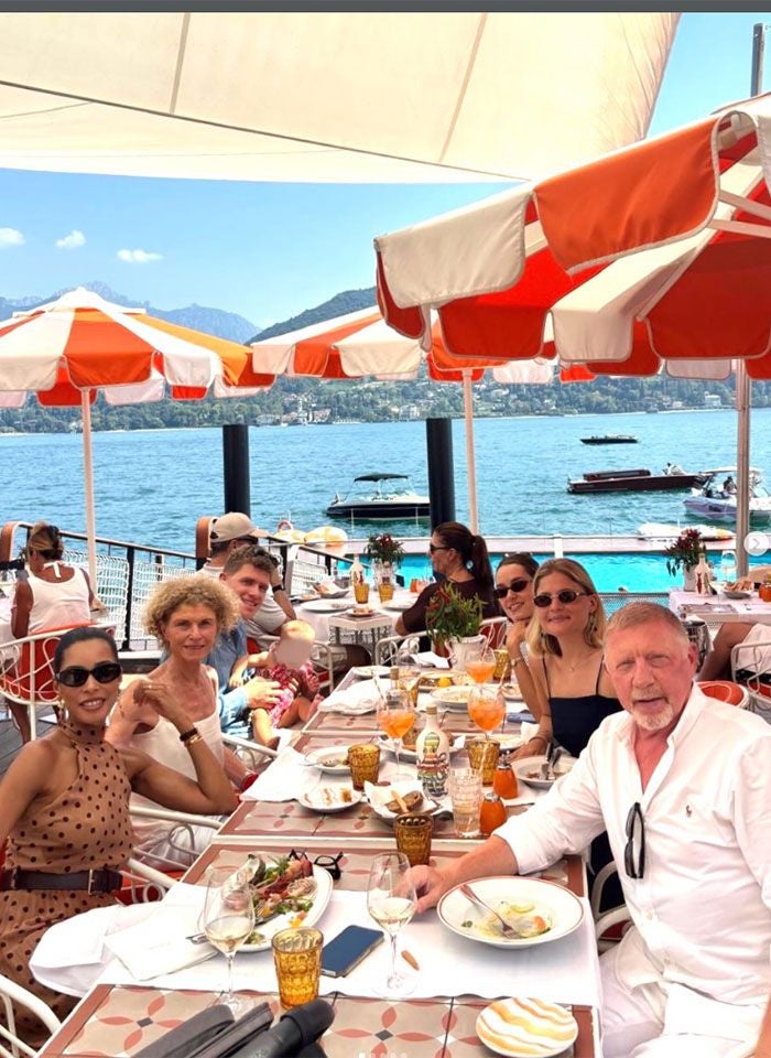 Menschen sitzen an einem gedeckten Tisch im Freien am See mit Sonnenschirmen im Hintergrund.