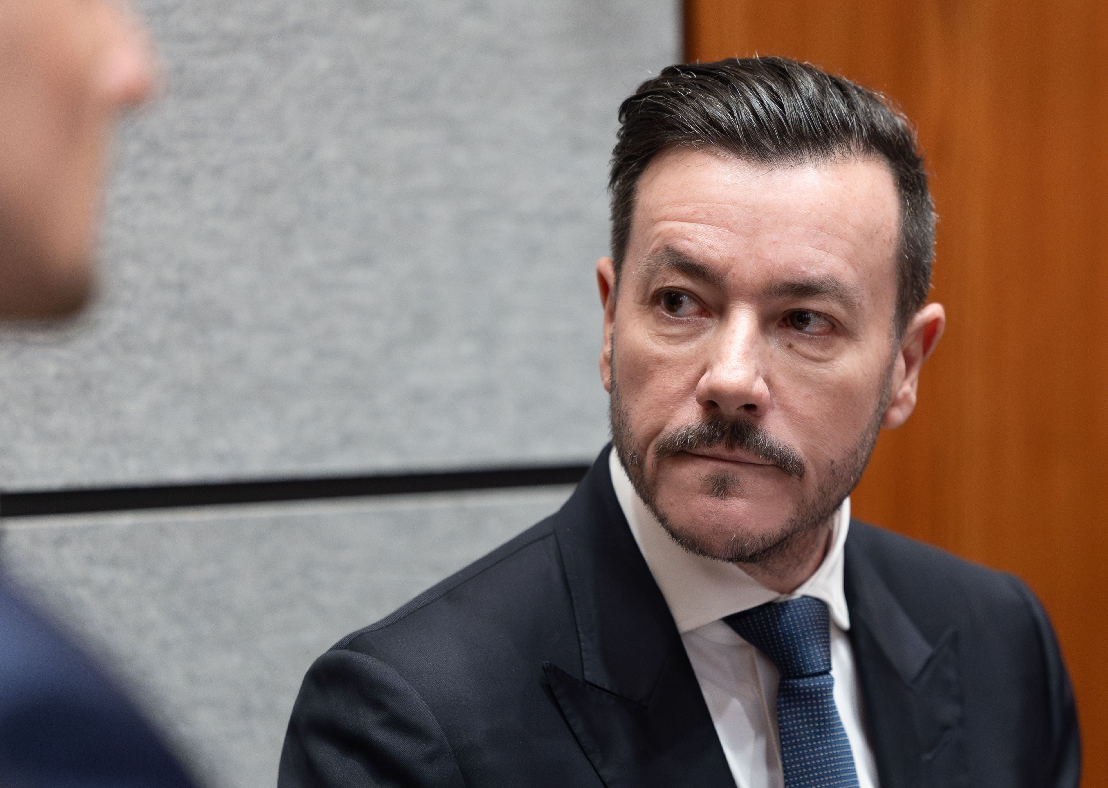 Mann im Anzug mit Bart schaut ernst nach rechts, grauer Hintergrund, Holzwand rechts.