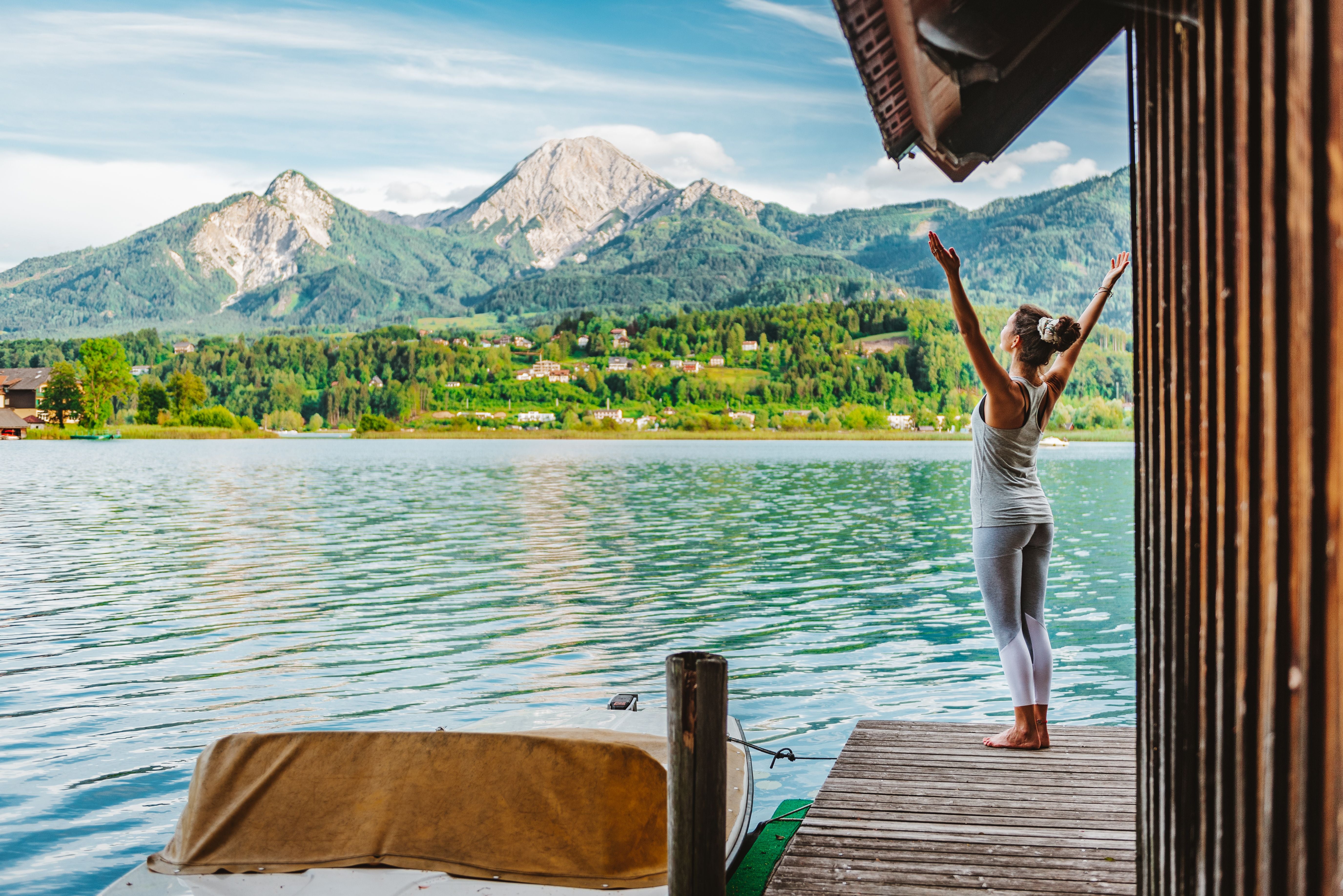 Frau macht Yoga auf einem Steg am Faaker See mit Blick auf Berge und grüne Landschaft.