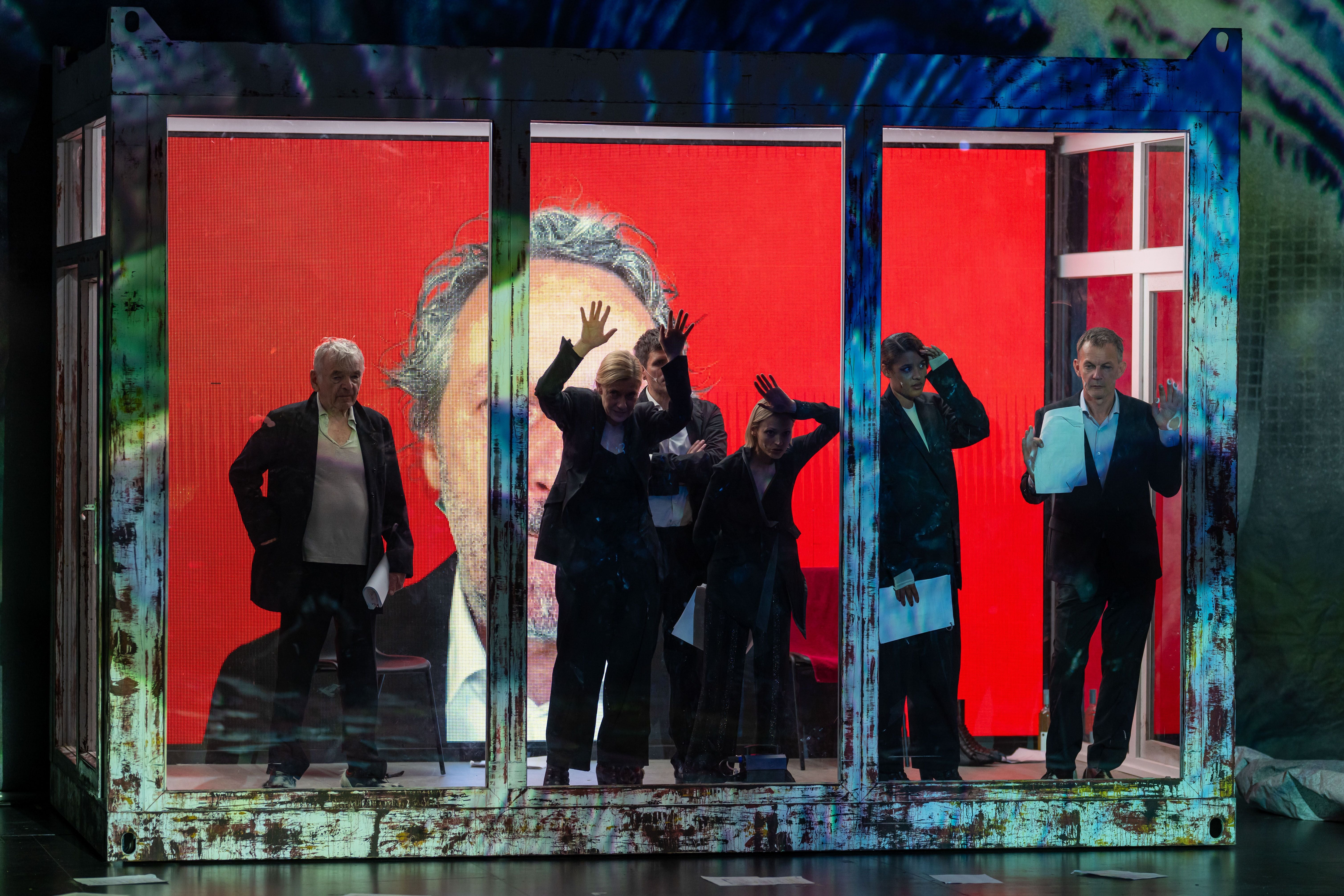 Sechs Schauspieler stehen in einem verrosteten Glaskasten vor rotem Hintergrund auf einer Theaterbühne.