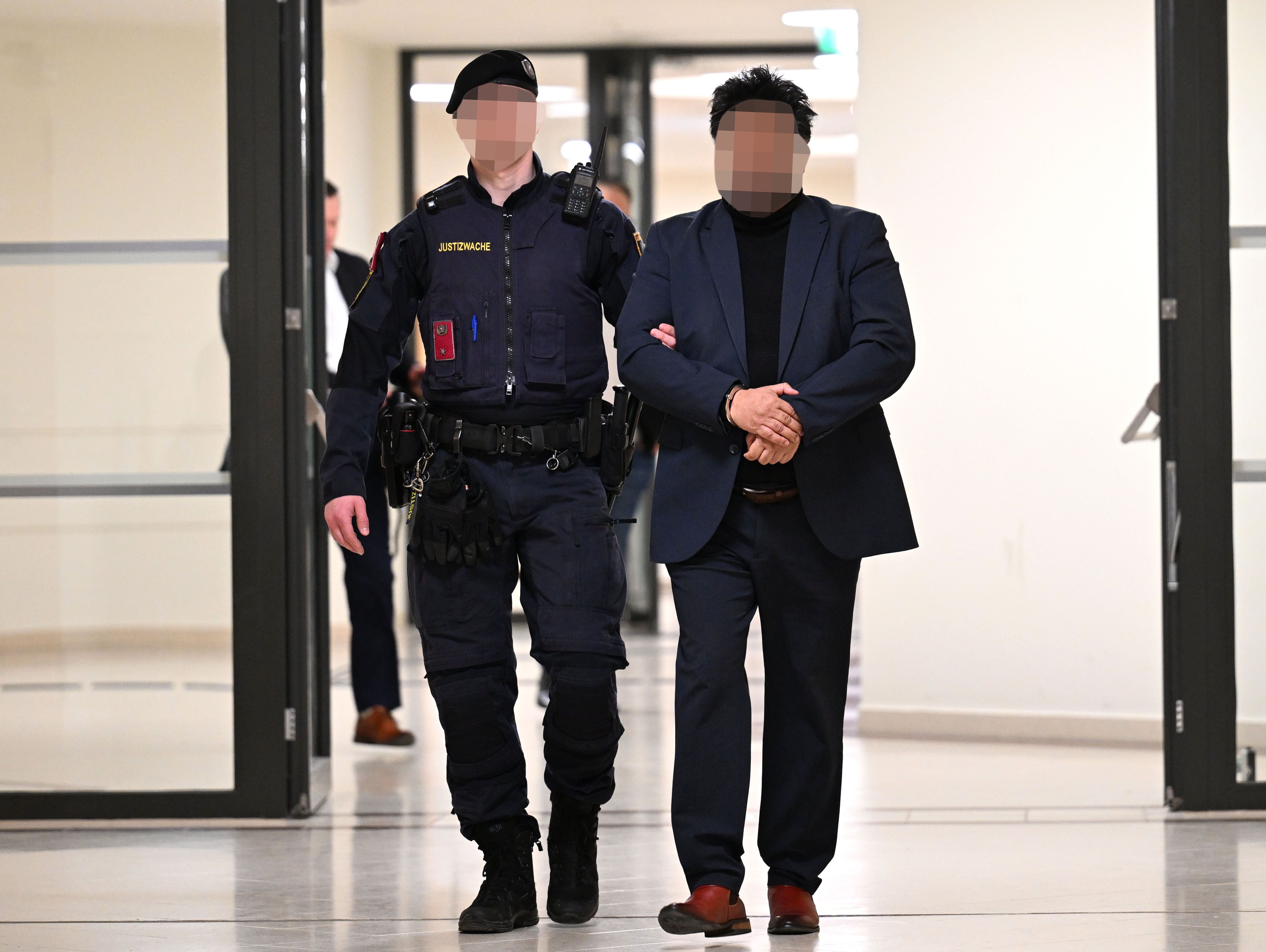Ein Polizist führt einen Mann in Anzug durch einen Flur, beide Gesichter sind verpixelt.