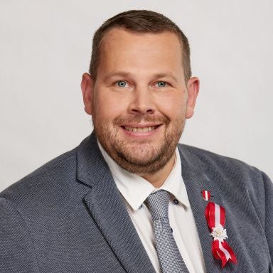 Ein Mann im Anzug mit Ordensschleife und Abzeichen, vor neutralem Hintergrund.