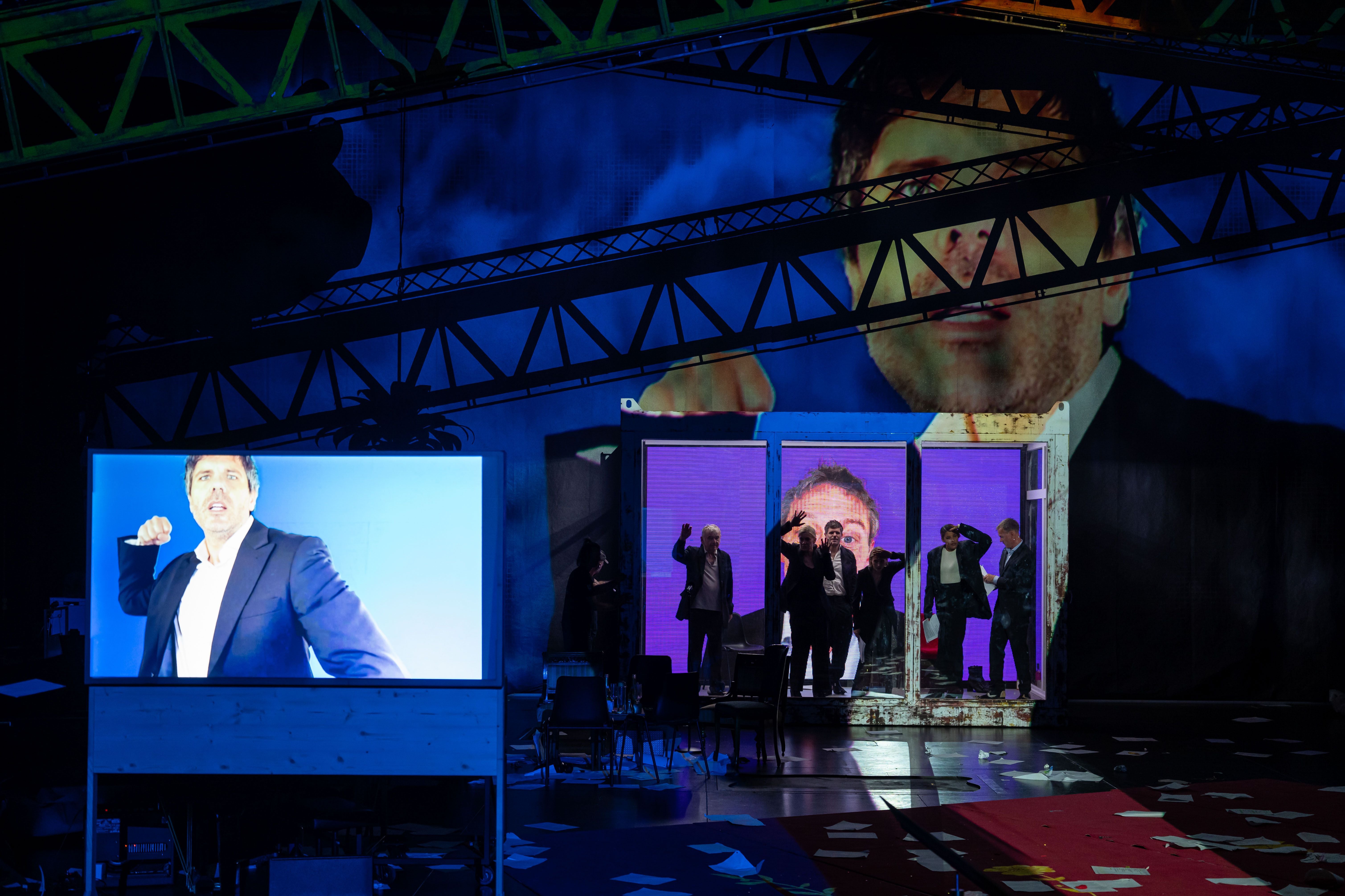 Theaterbühne mit großem Porträt, Bildschirm und mehreren Schauspielern in einer Glaskabine.