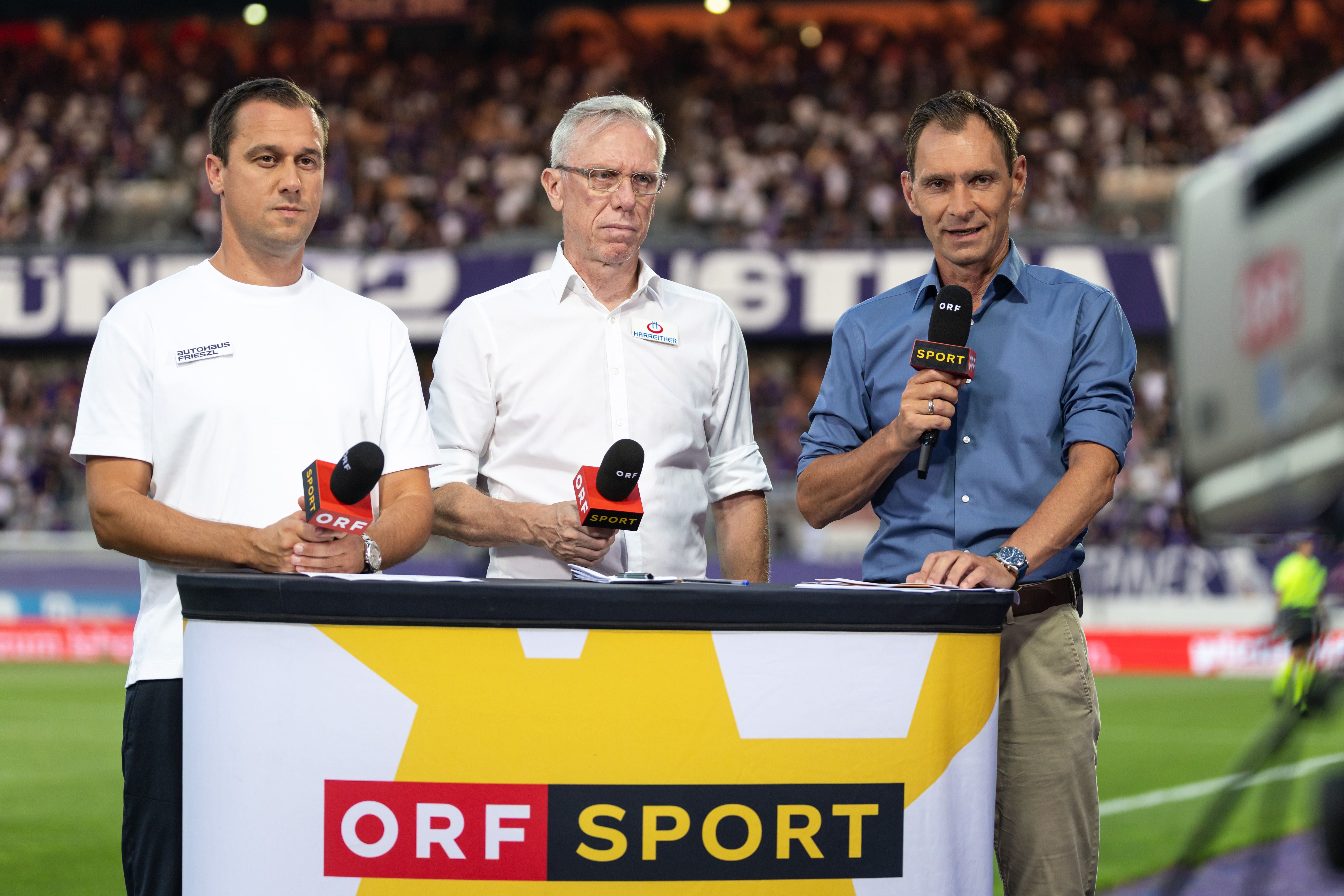 Drei Männer stehen mit Mikrofonen bei einer ORF Sport TV-Übertragung im Fußballstadion.