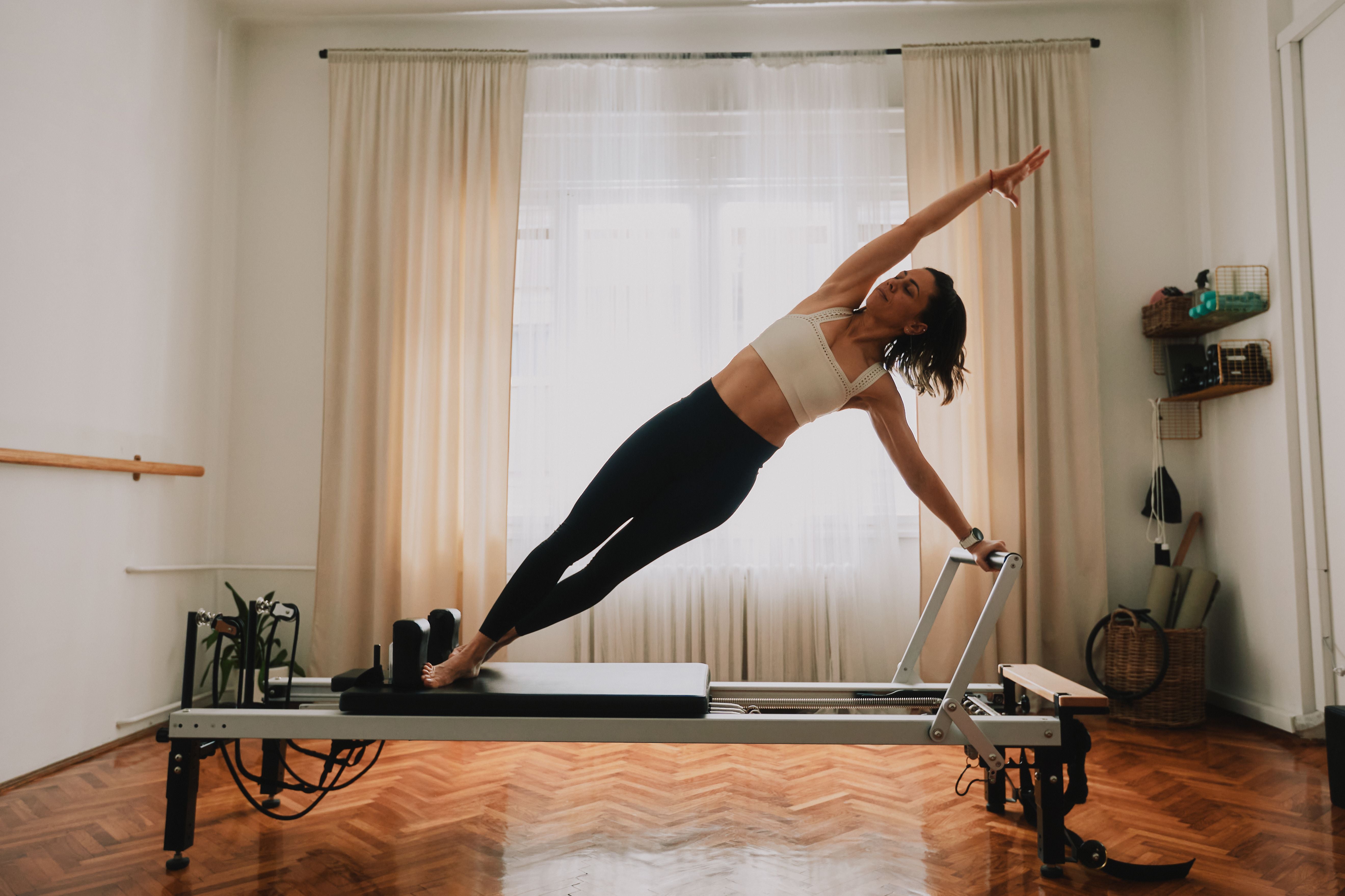Frau macht Pilates-Übung auf einem Reformer-Gerät im sonnigen Wohnzimmer.