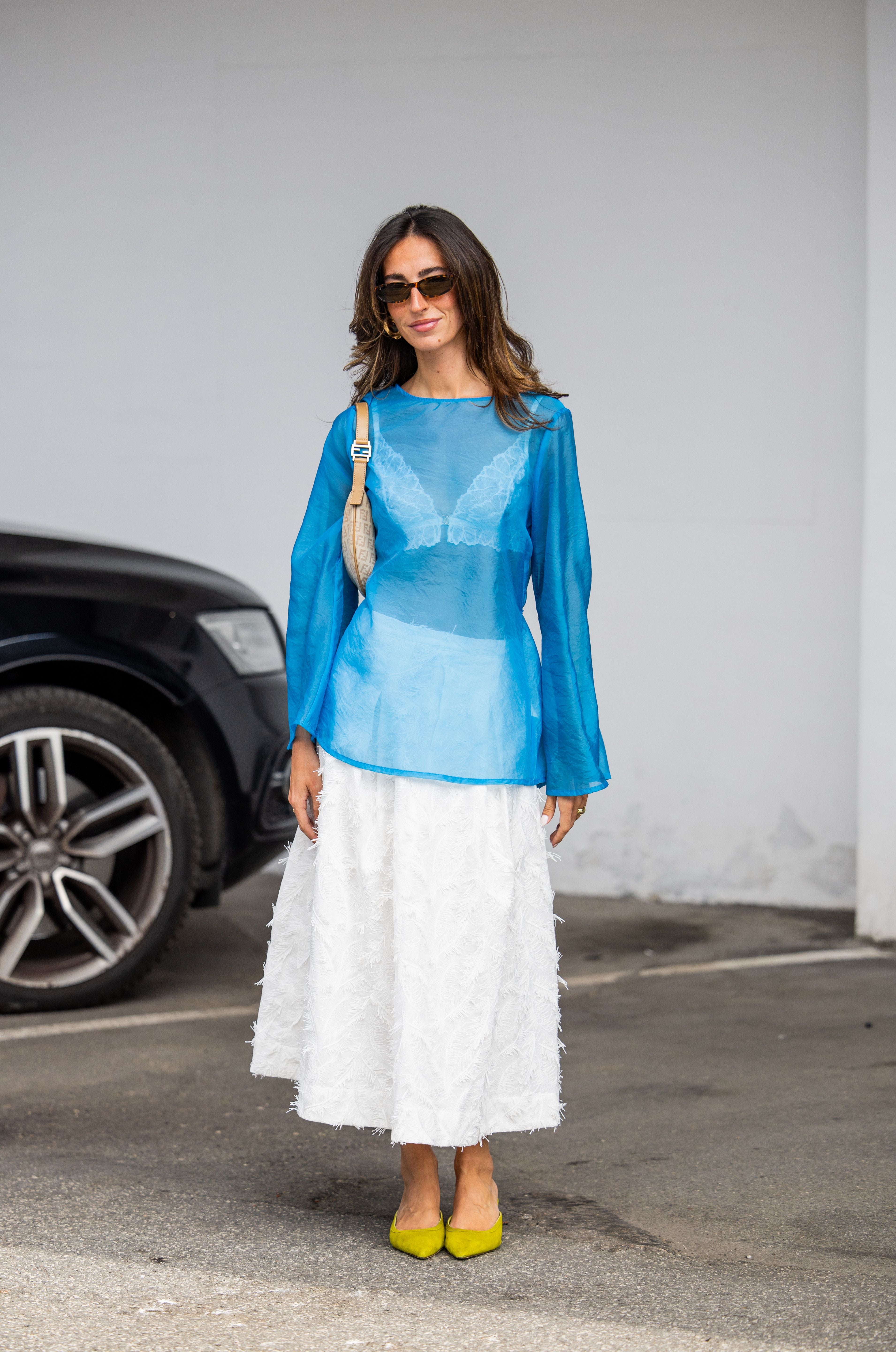 Frau mit blauem Transparent-Top, weißem Rock, gelben Schuhen und beiger Tasche auf der Straße.