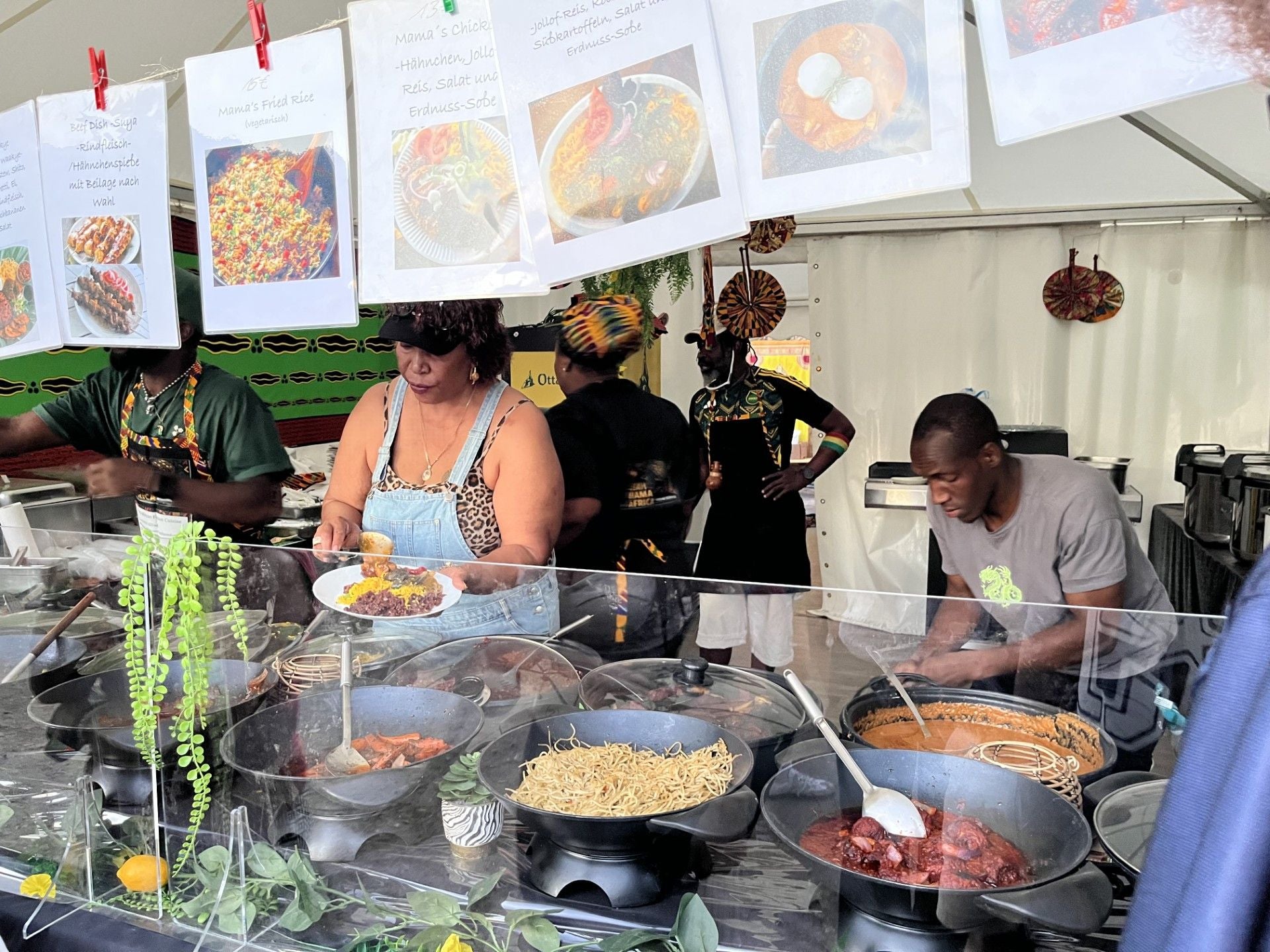 Menschen servieren afrikanisches Essen an einem Food-Stand auf einem Festival, verschiedene Gerichte sichtbar.