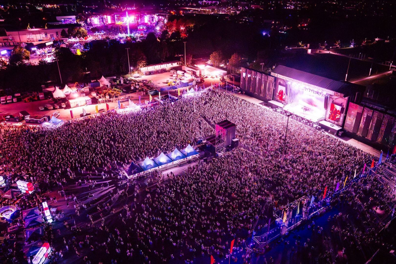 Luftaufnahme eines riesigen Musikfestivals bei Nacht mit bunter Bühnenbeleuchtung und Menschenmenge.