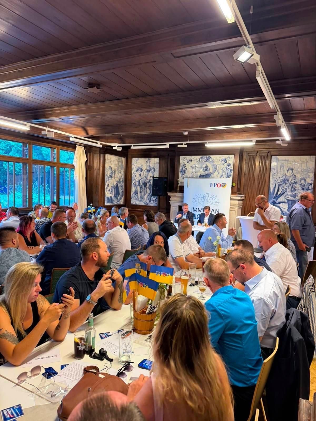 Menschen sitzen an langen Tischen in einem holzgetäfelten Saal bei einer Veranstaltung der FPÖ.