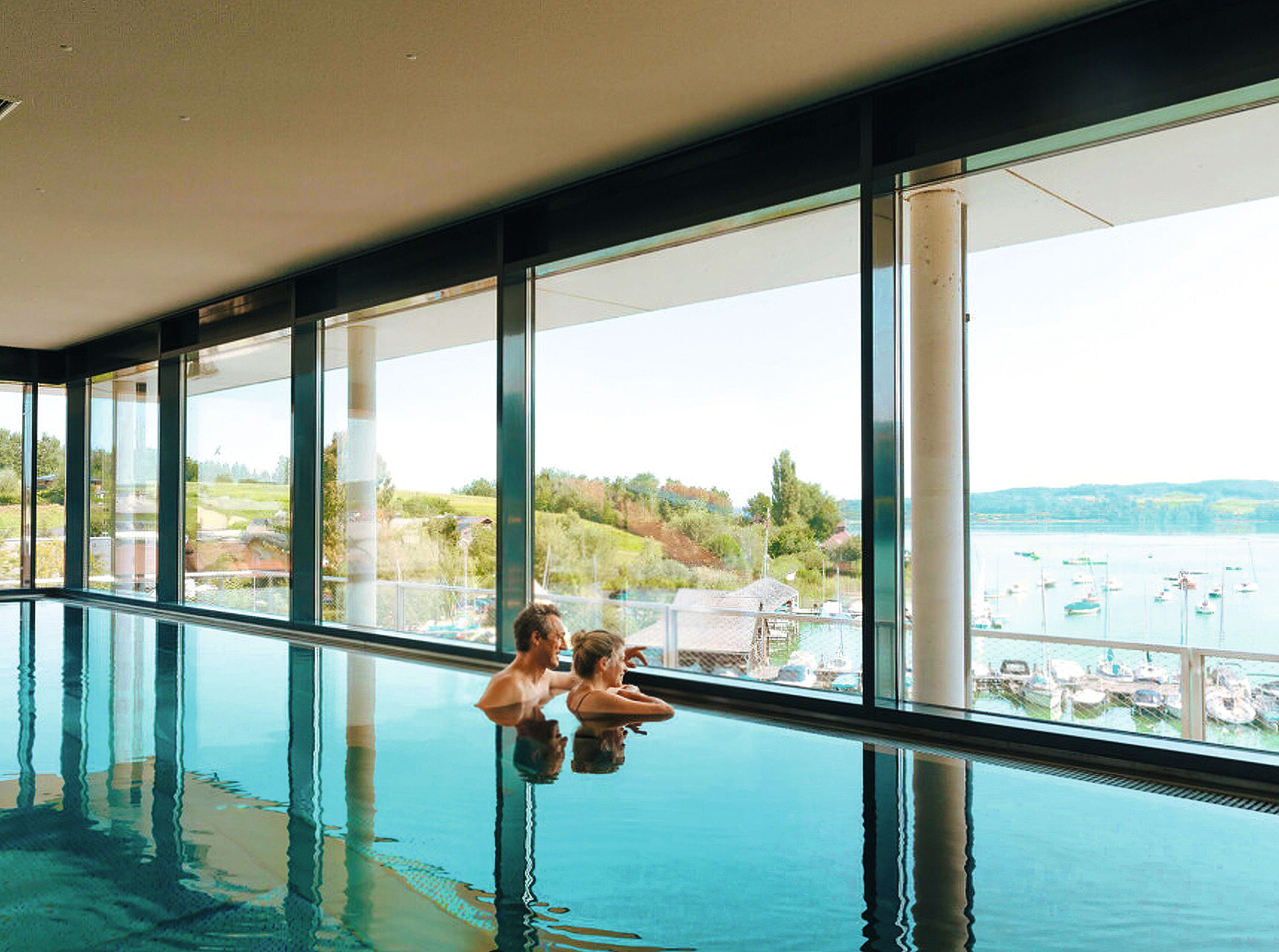 Zwei Personen entspannen in einem Innenpool mit Blick auf einen See und Segelboote durch große Fenster.