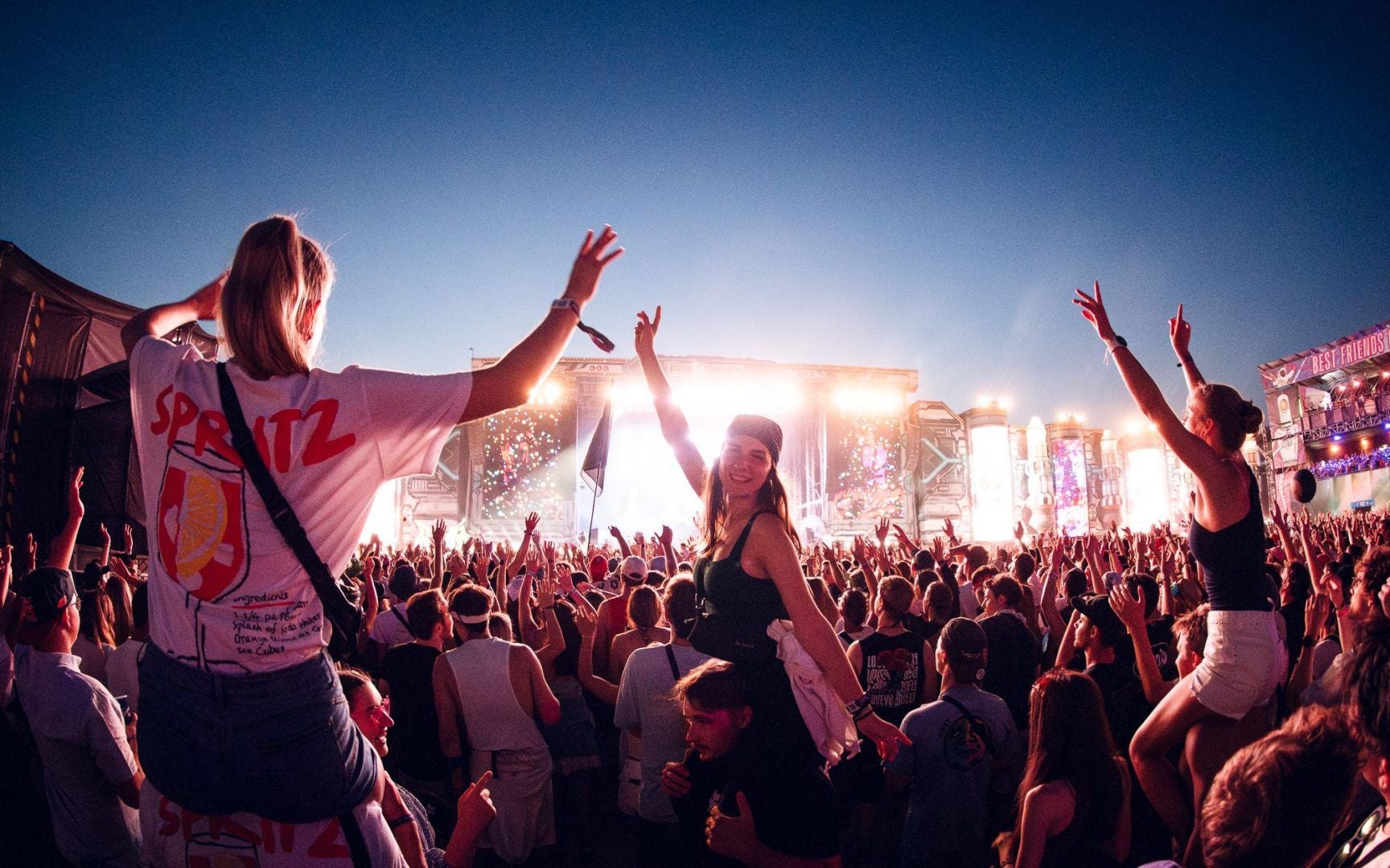 Junge Menschen feiern und tanzen bei Sonnenuntergang vor einer großen Musikfestival-Bühne.