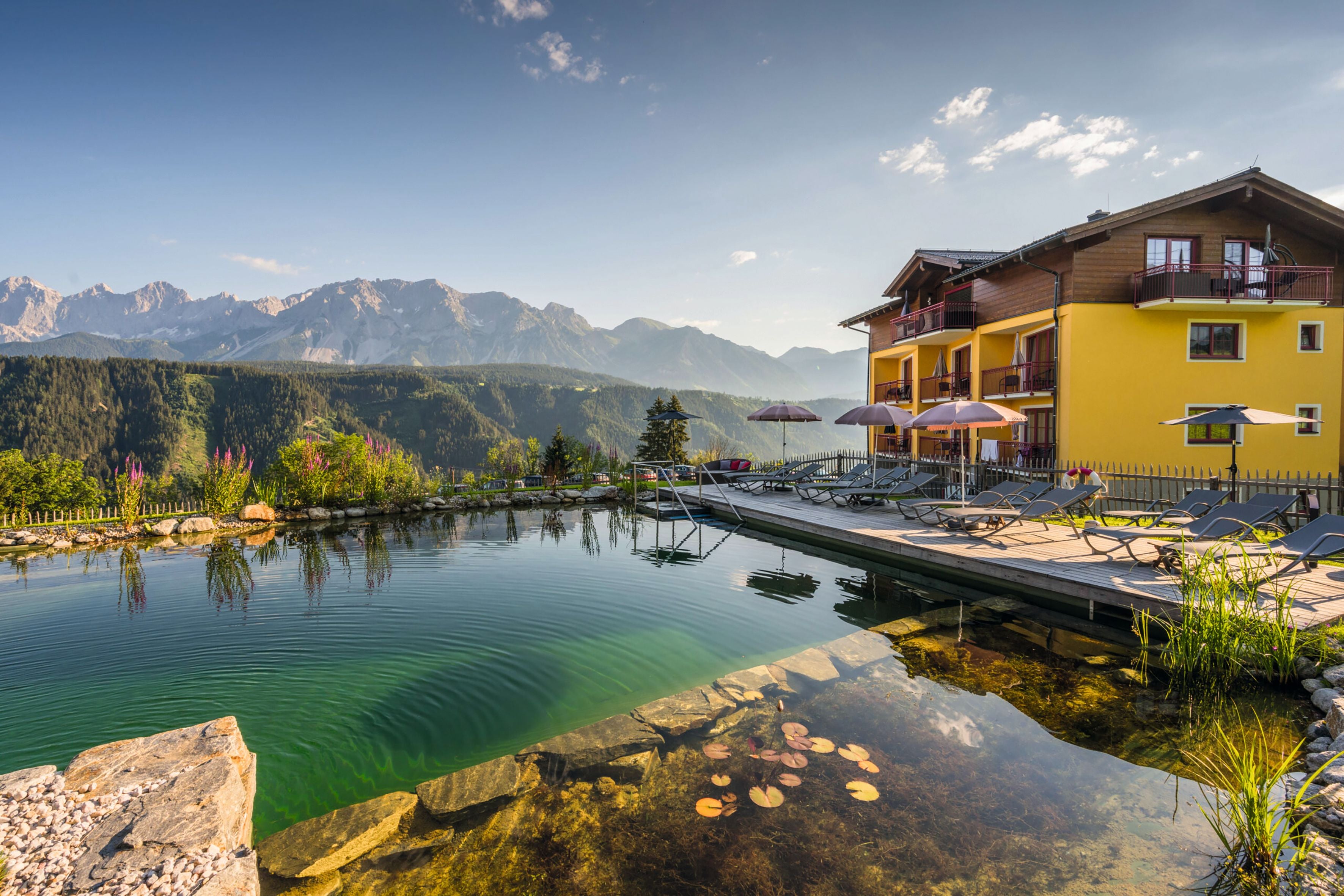 Hotel mit Naturteich, Liegestühlen und Sonnenschirmen, im Hintergrund Berge und Wälder bei sonnigem Wetter.