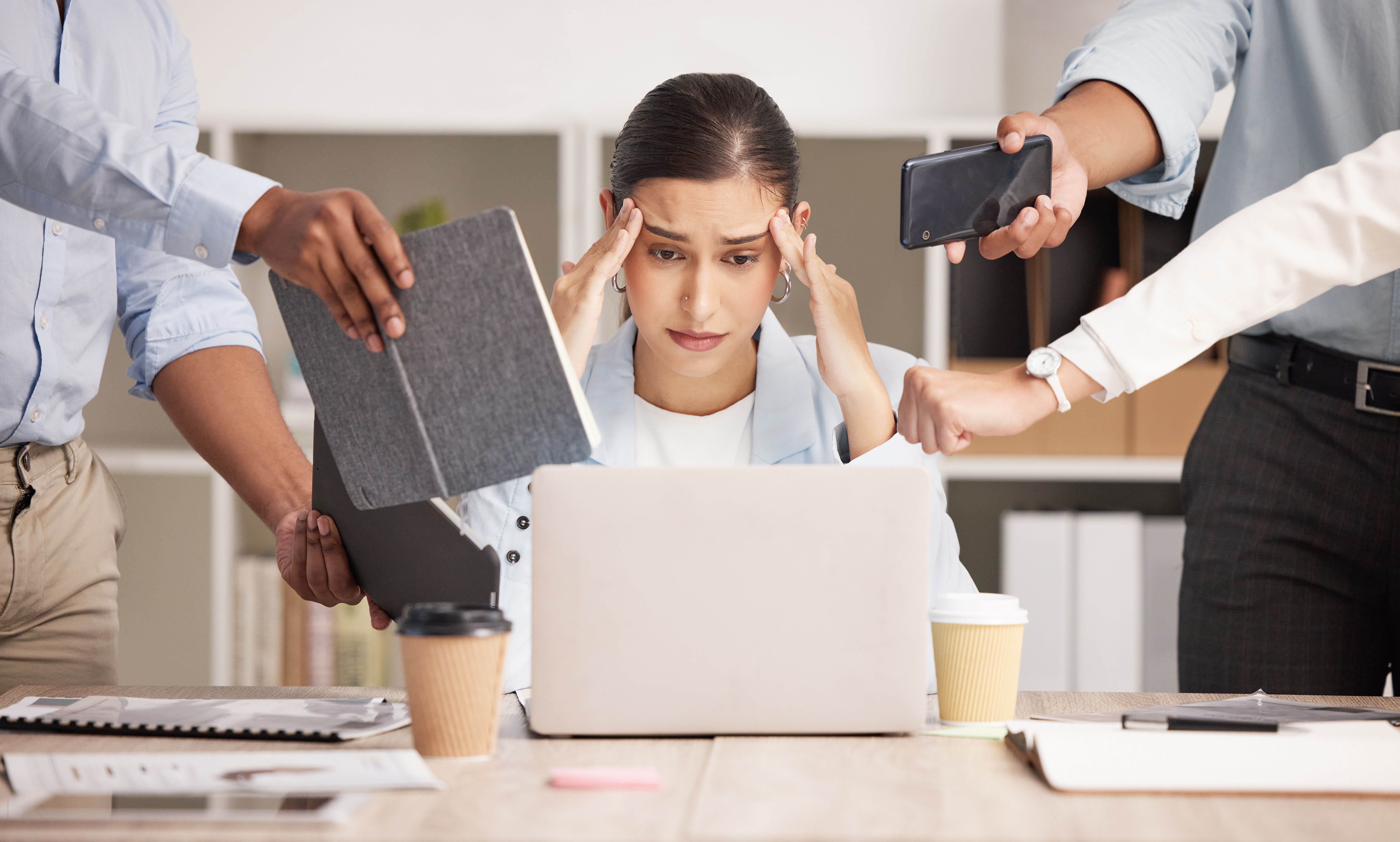 Gestresste Frau sitzt im Büro vor Laptop, umgeben von Forderungen und wirkt überfordert.