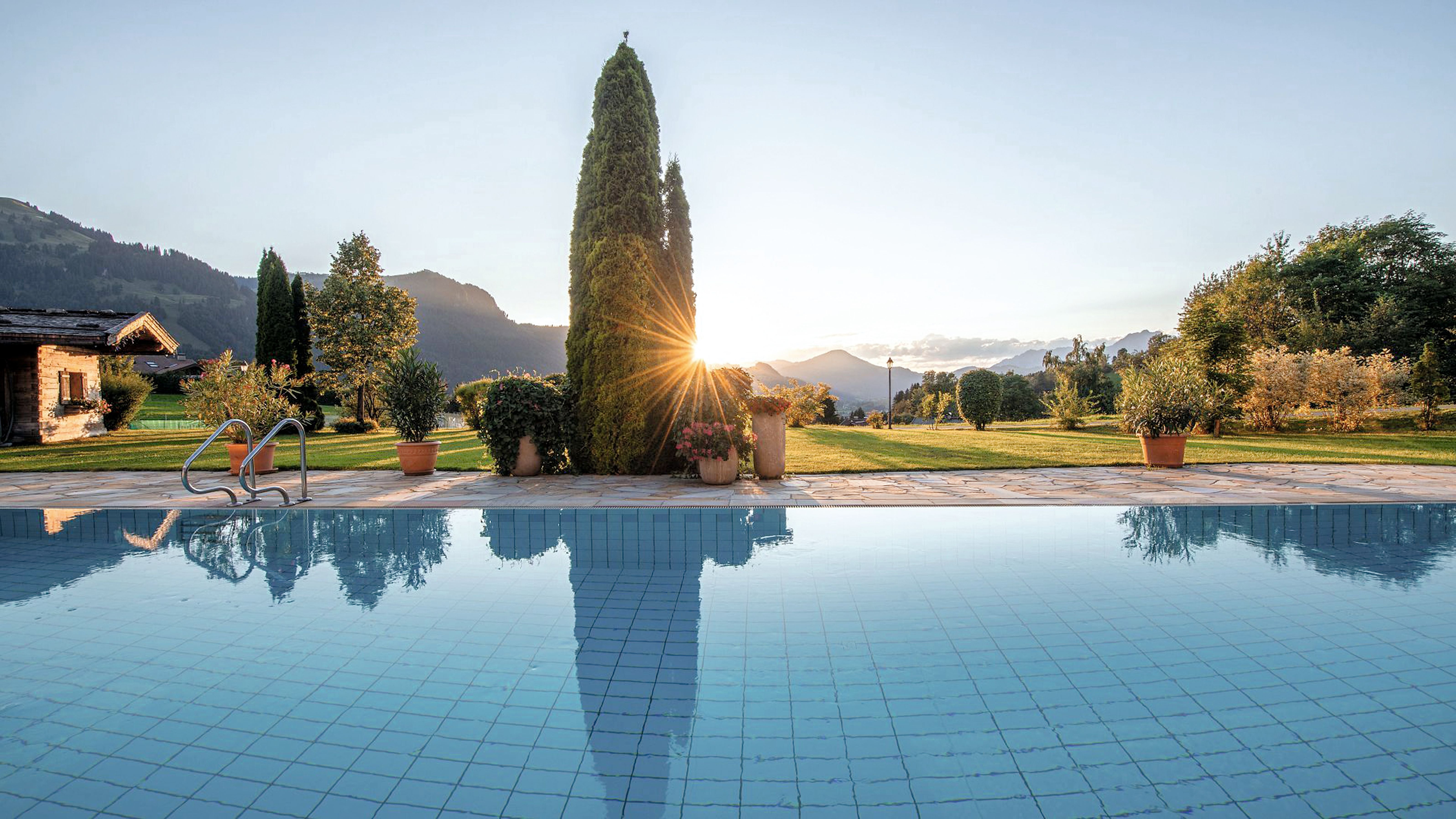 Swimmingpool mit klarer Wasseroberfläche, Sonnenaufgang hinter Bäumen und Bergen im Hintergrund.