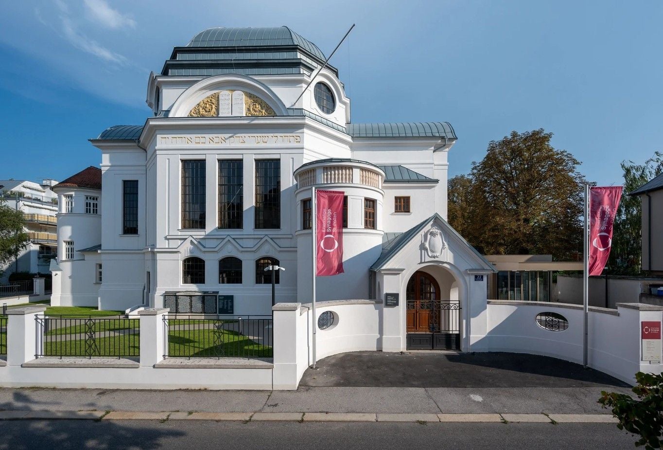 Die St. Pöltner Synagoge mit weißer Fassade, großen Fenstern und roten Fahnen davor.