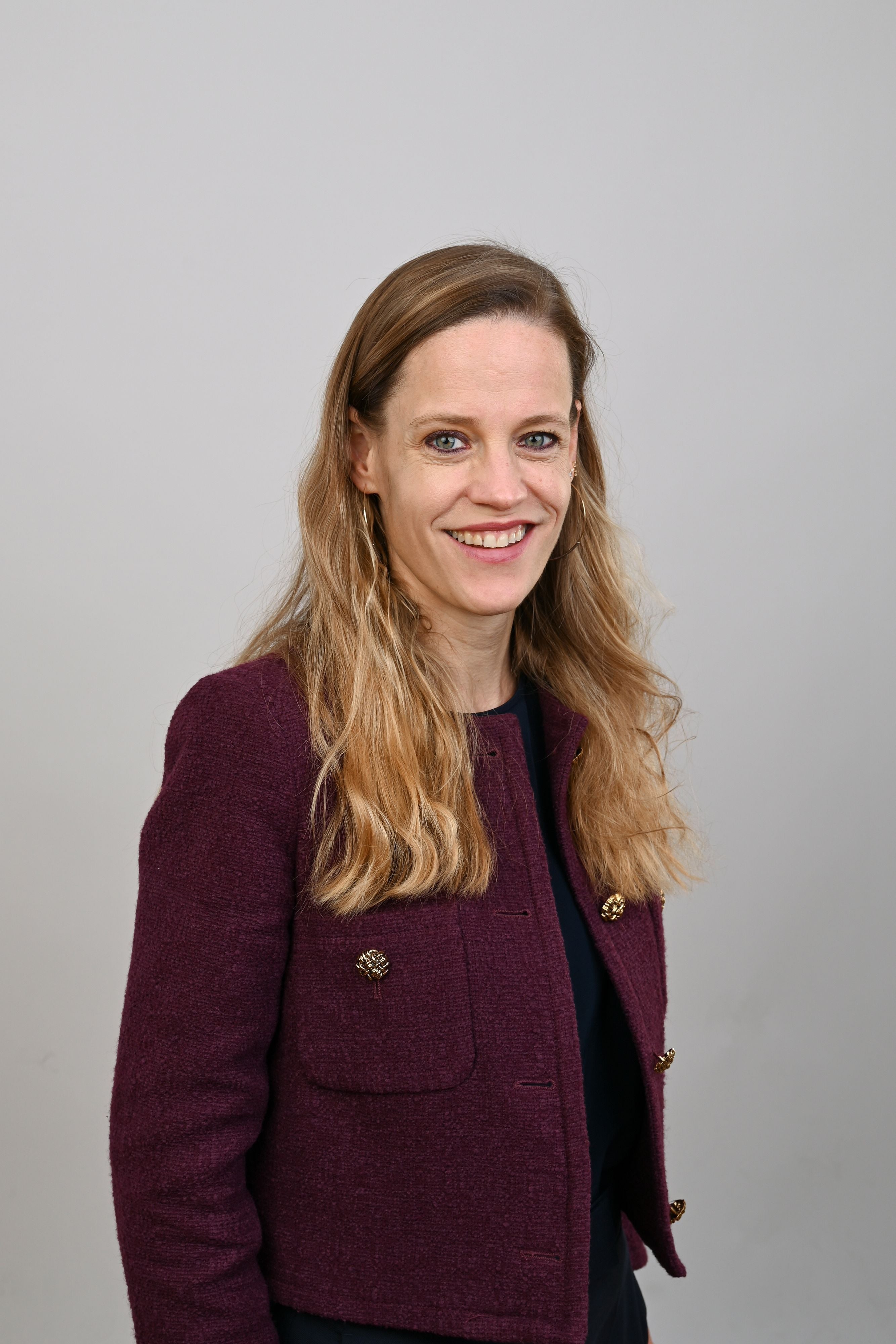 Eine Frau mit langen blonden Haaren trägt eine bordeauxrote Jacke und lächelt in die Kamera.