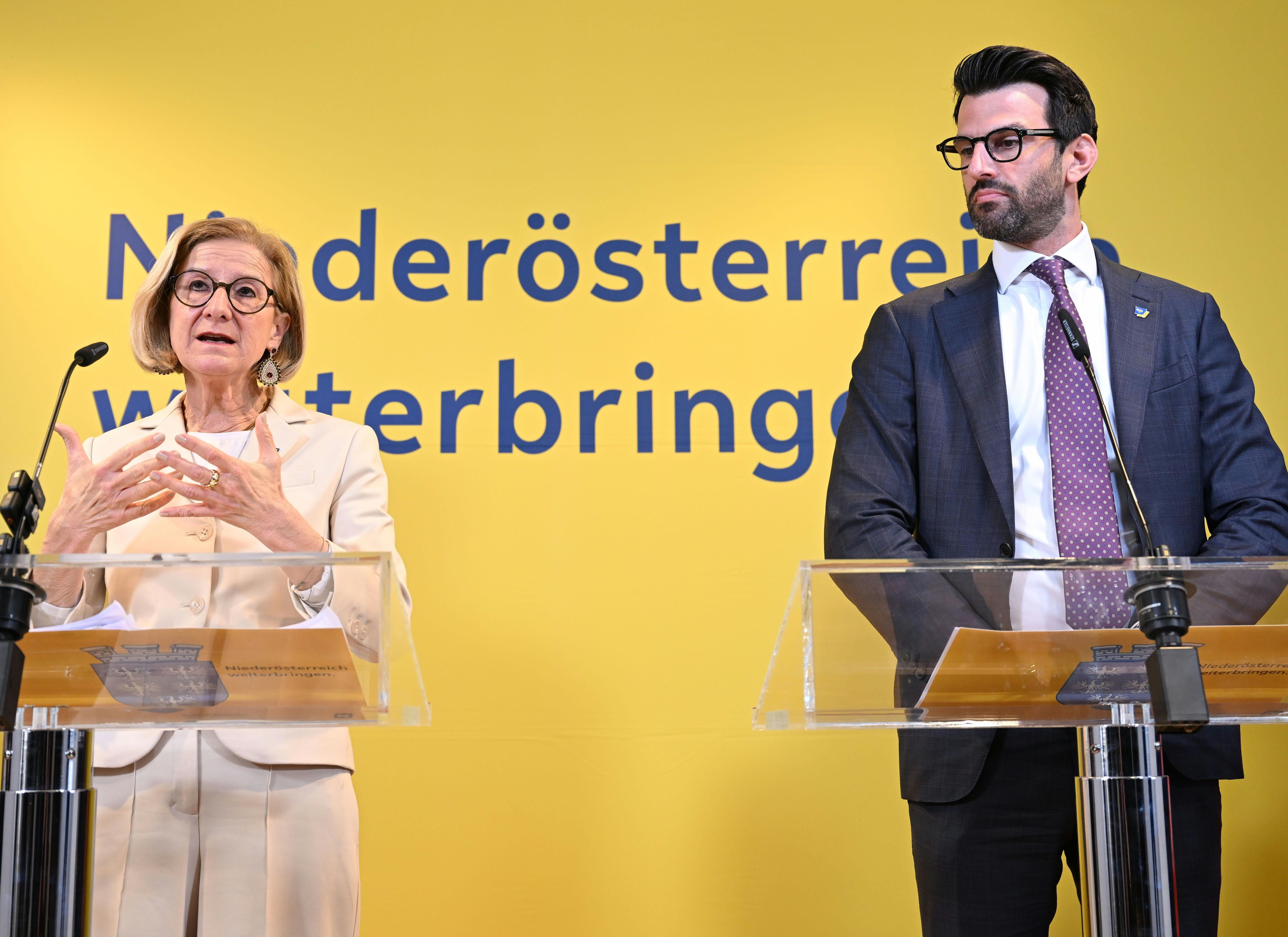 Zwei Politiker sprechen bei einer Pressekonferenz vor gelbem Hintergrund mit Mikrofonen und Rednerpulten.
