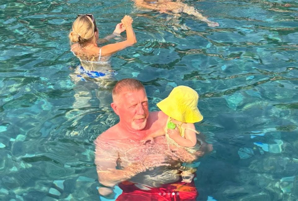 Ein älterer Mann hält ein Kleinkind mit gelbem Sonnenhut im Schwimmbecken.