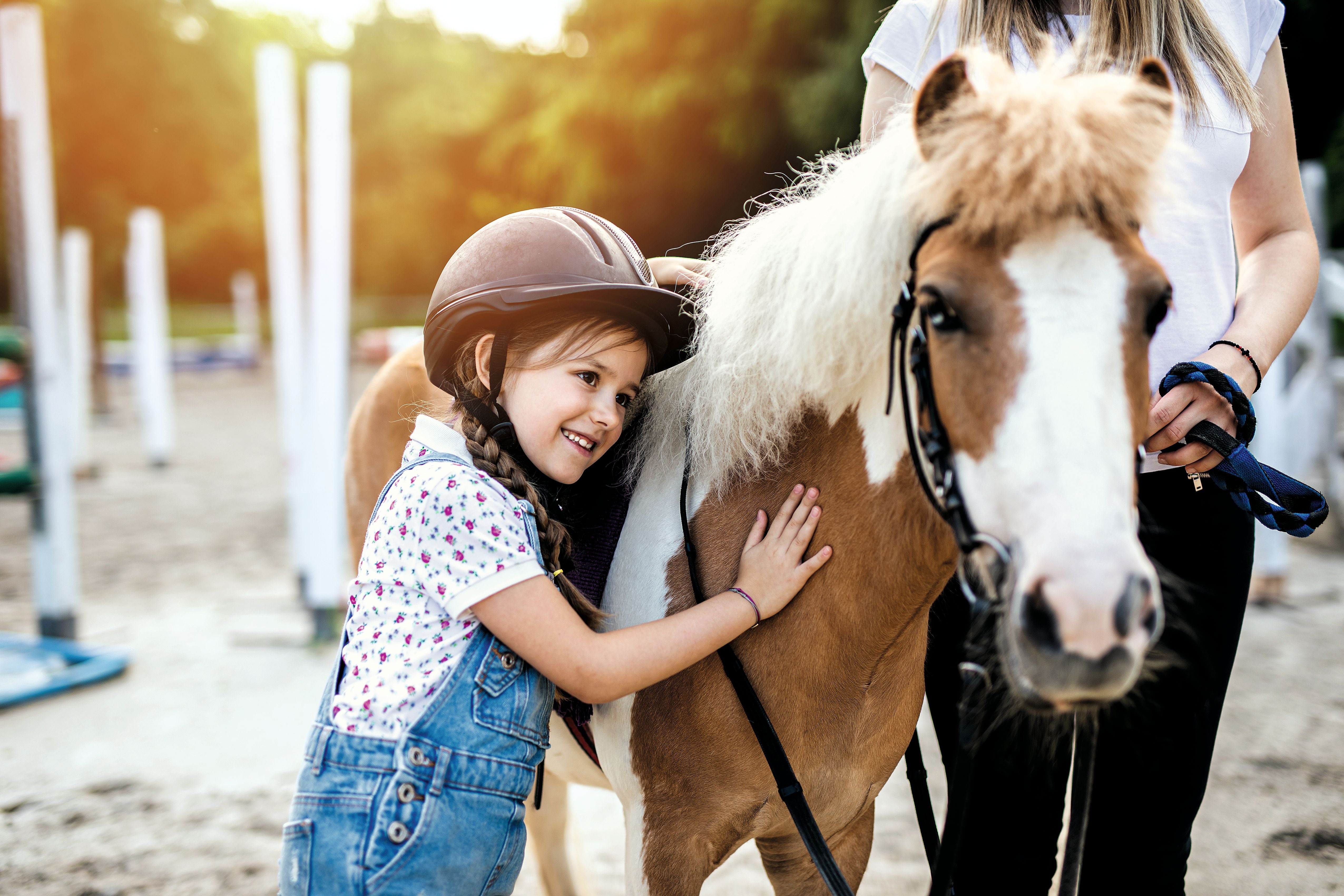 Mädchen mit Reithelm umarmt lächelnd ein Pony auf einem Reitplatz im Freien.