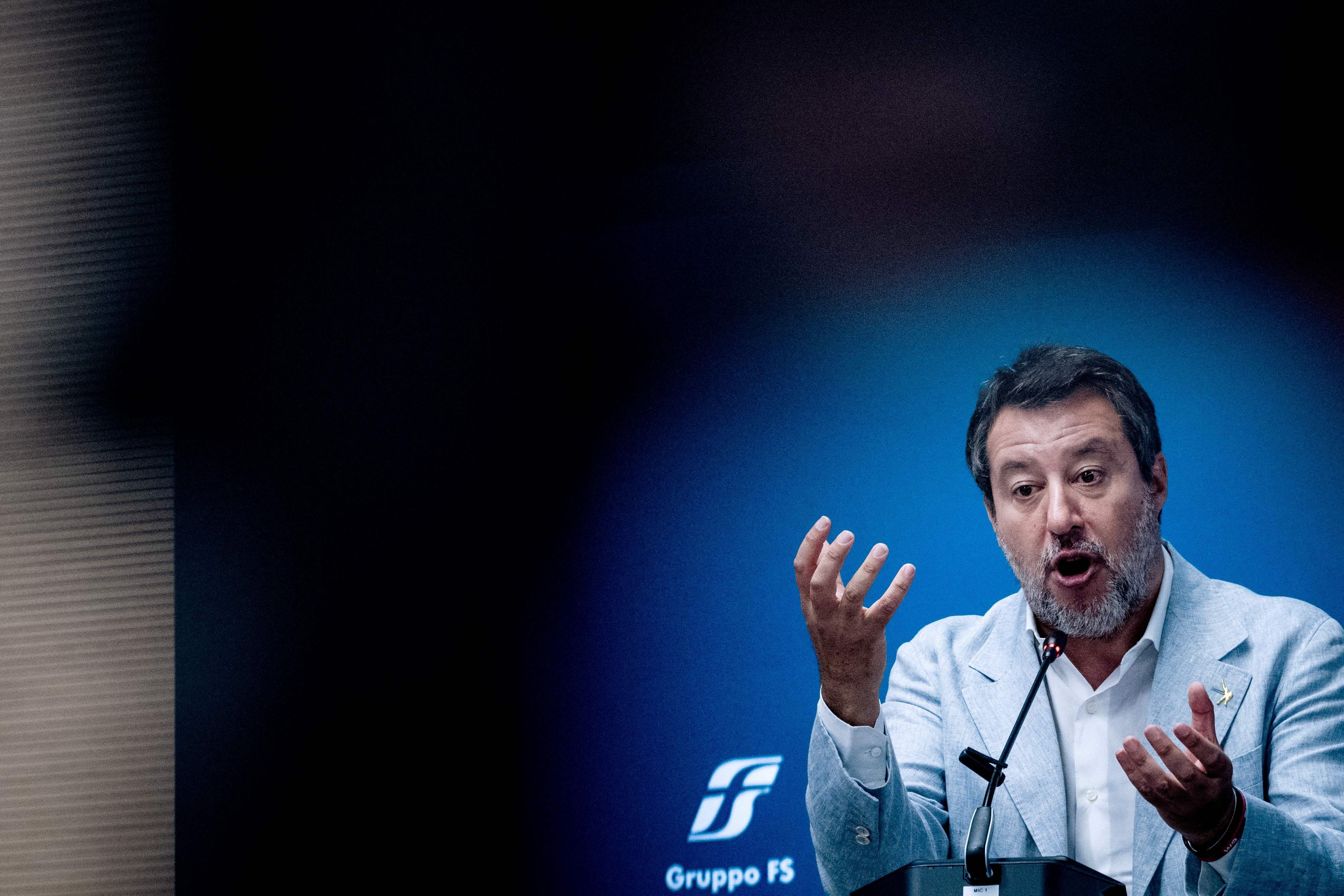 Ein Mann im Anzug spricht gestikulierend bei einer Pressekonferenz vor blauem Hintergrund.