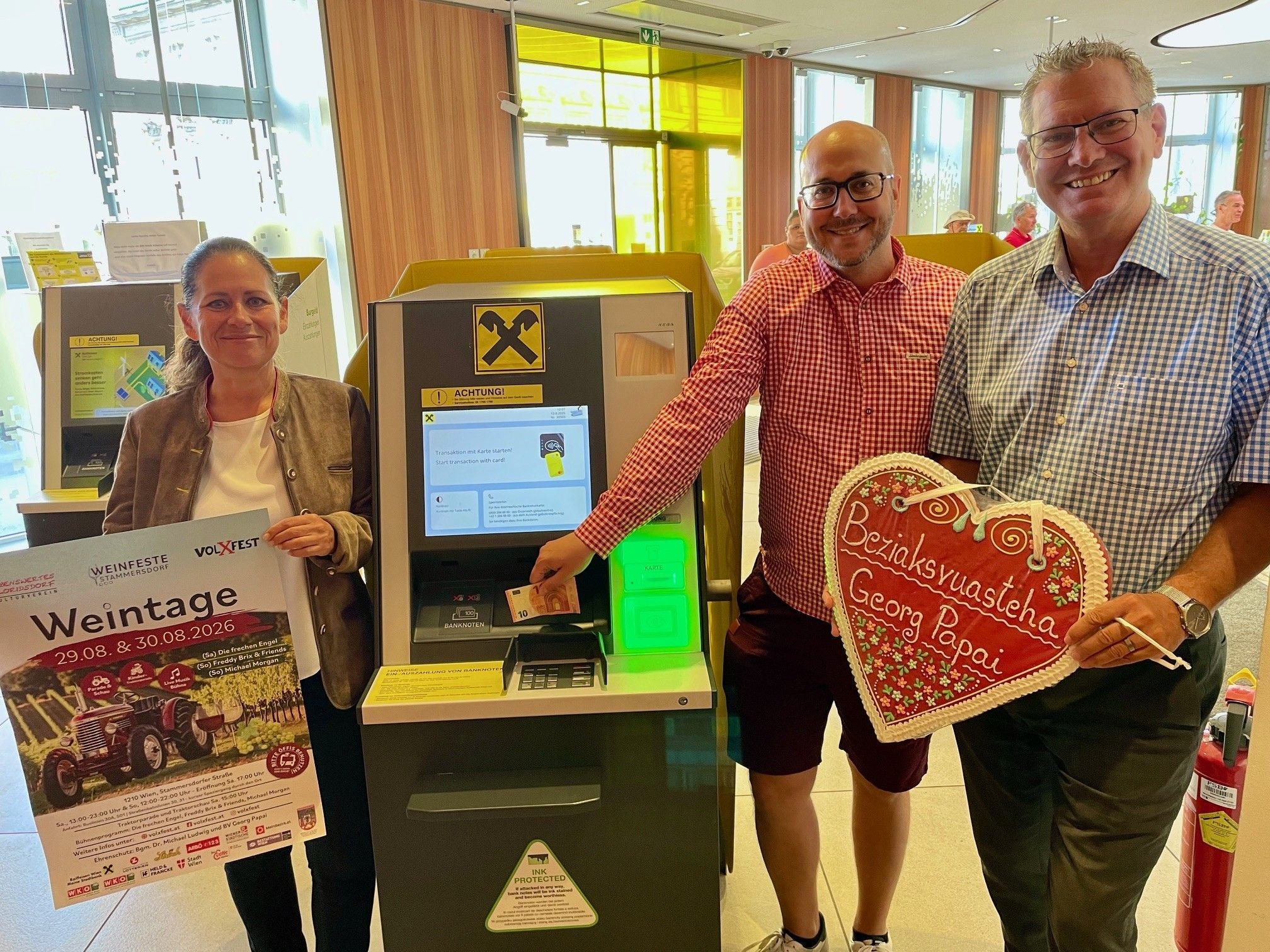 Drei Personen stehen an einem Bankautomaten; eine hält ein Poster, die andere ein Lebkuchenherz.