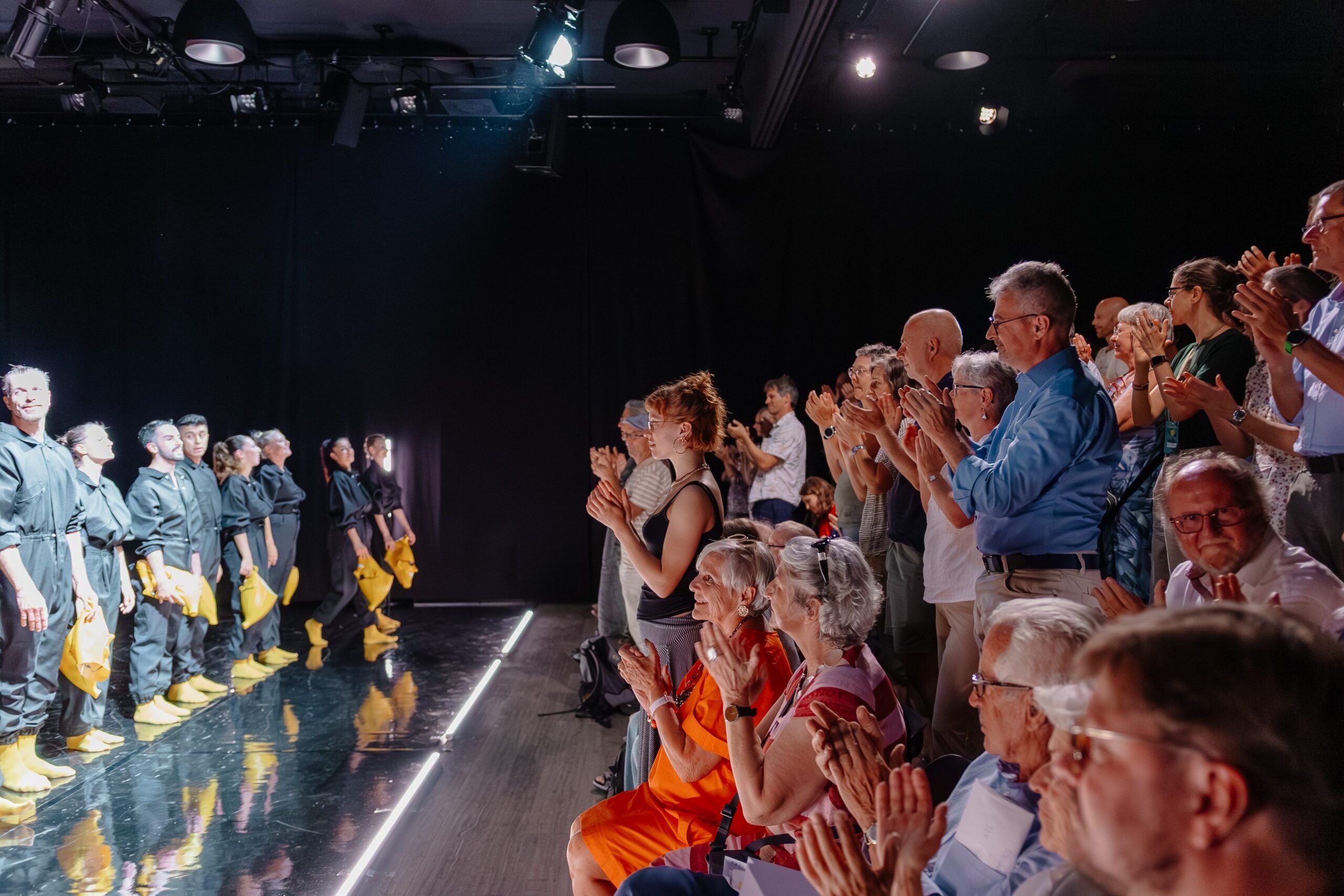 Das Publikum applaudiert stehend einer Gruppe von Künstlern auf einer Bühne in einem Theater.