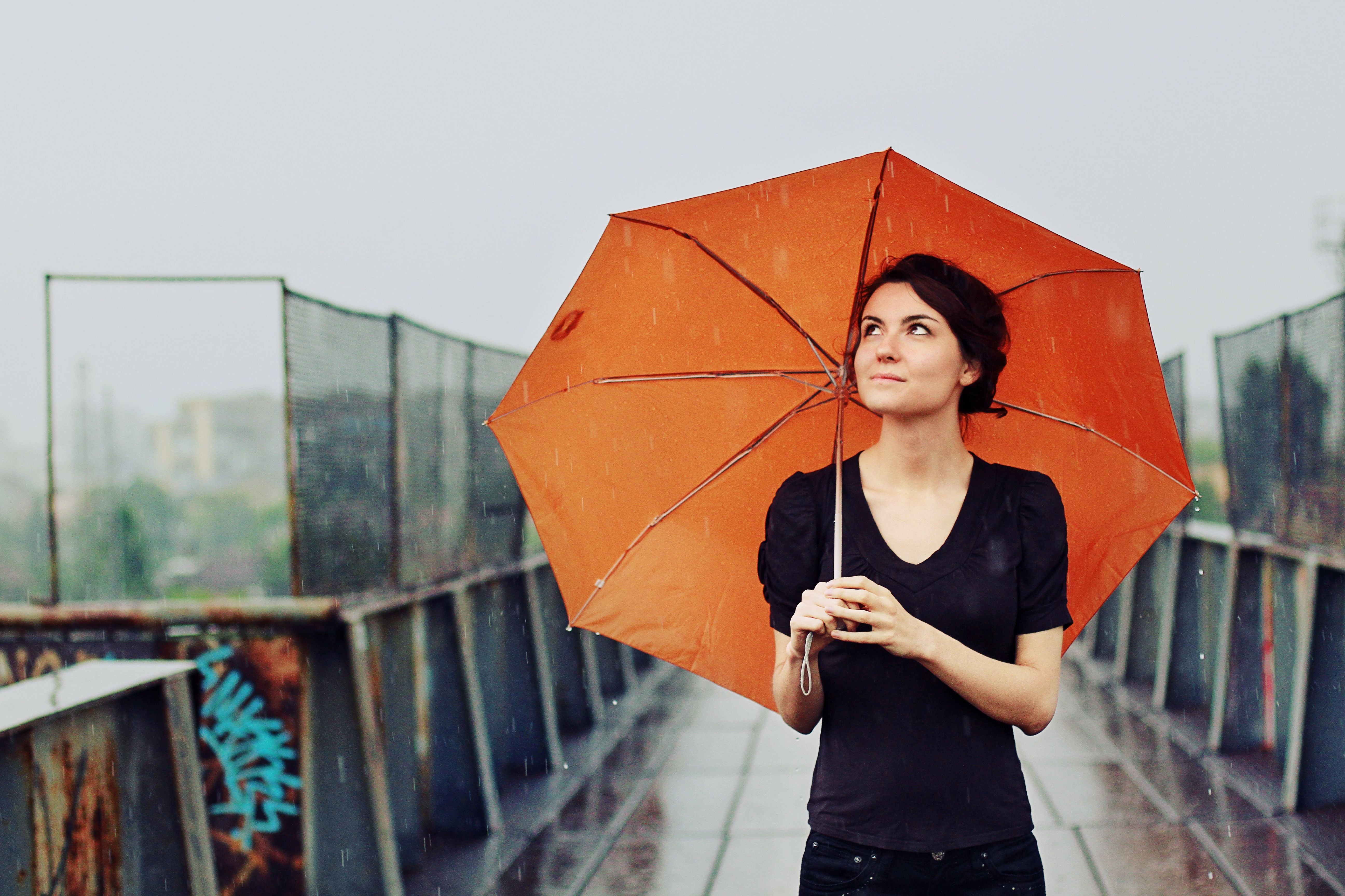 Junge Frau mit orangefarbenem Regenschirm steht lächelnd im Regen und blickt nach oben.