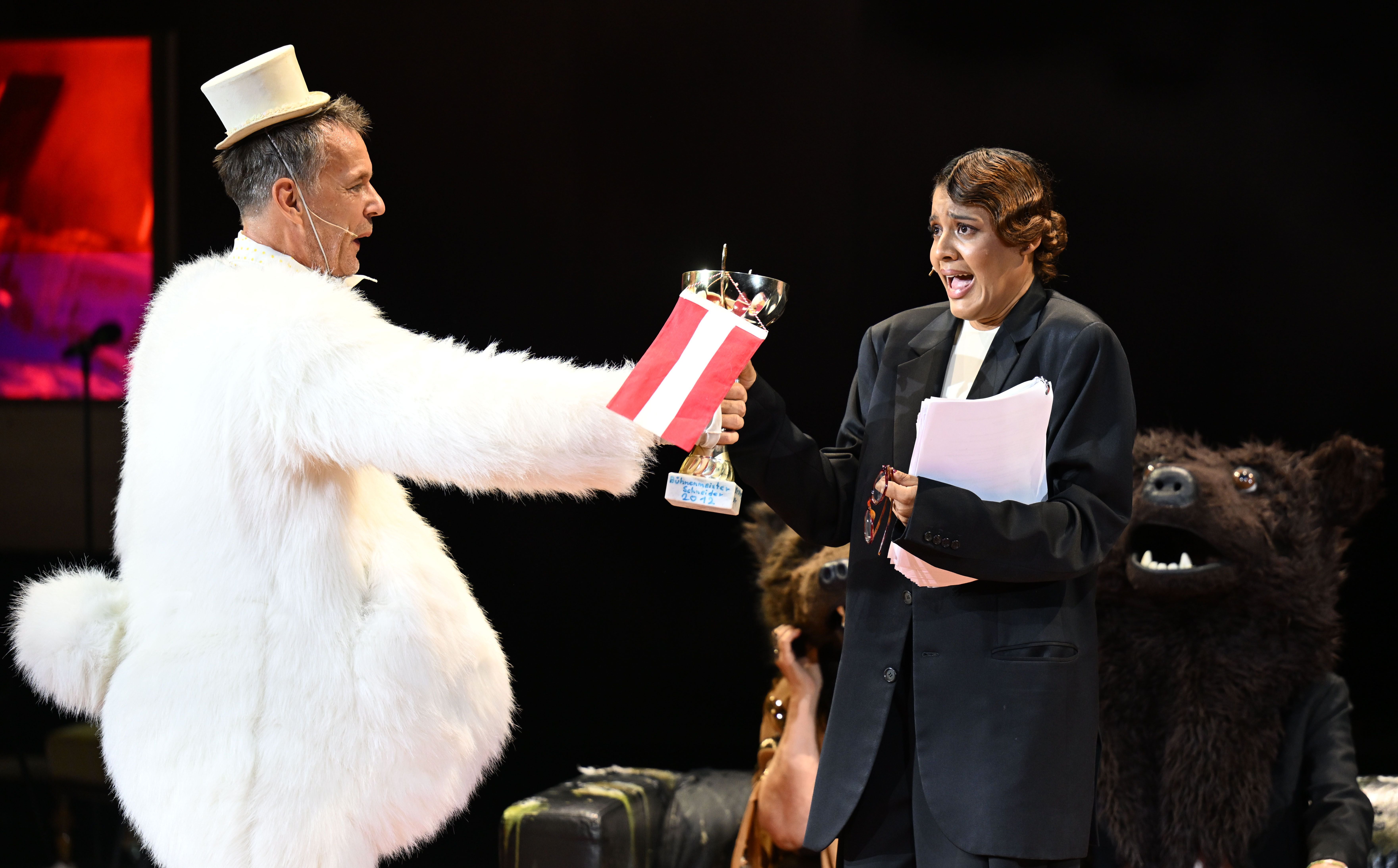 Ein Mann im weißen Kostüm mit Hut überreicht einer Frau im Anzug einen Pokal auf einer Theaterbühne.
