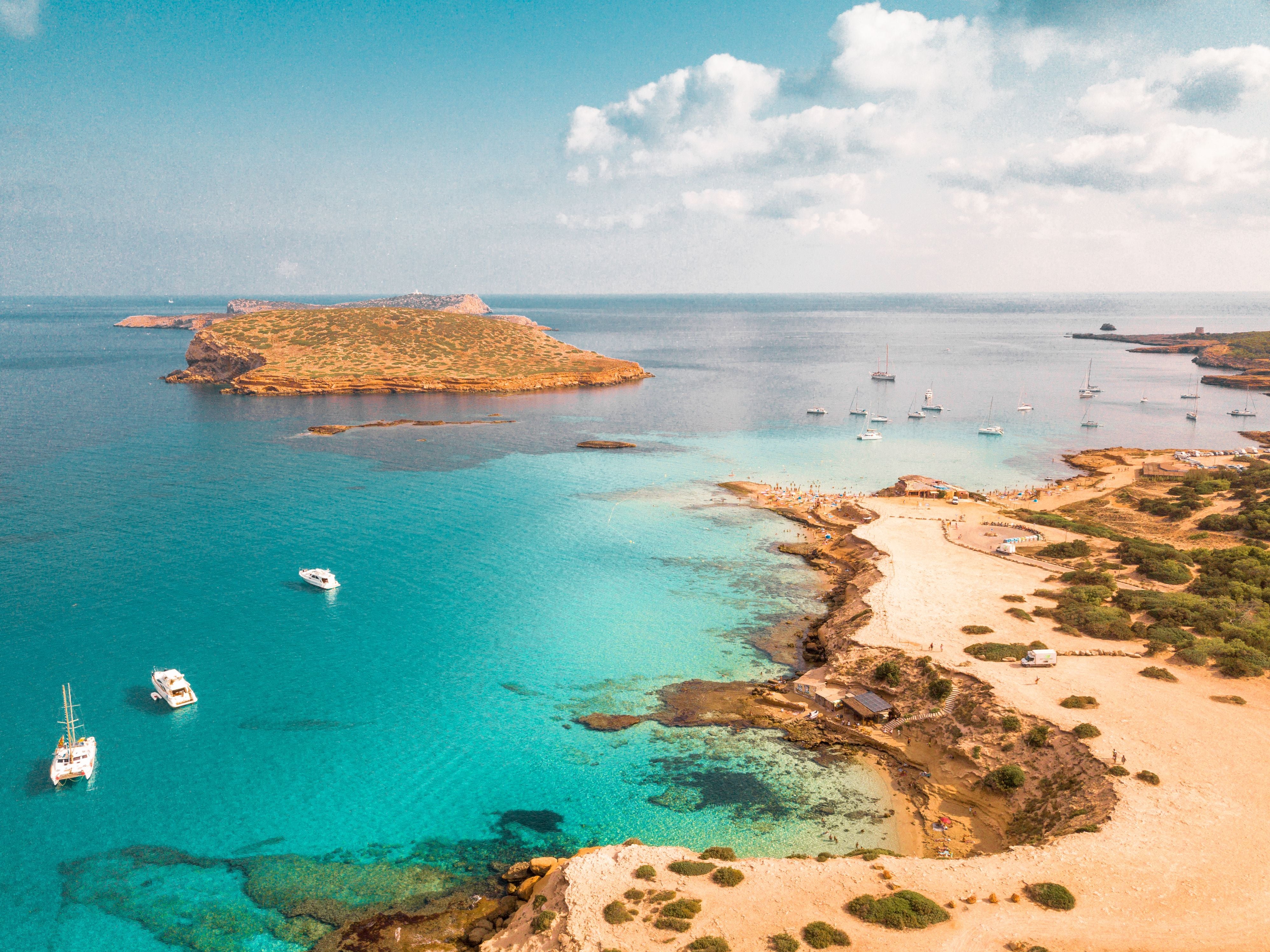 Luftaufnahme einer Küste auf Ibiza mit türkisblauem Wasser, Sandstrand und mehreren Booten im Meer.