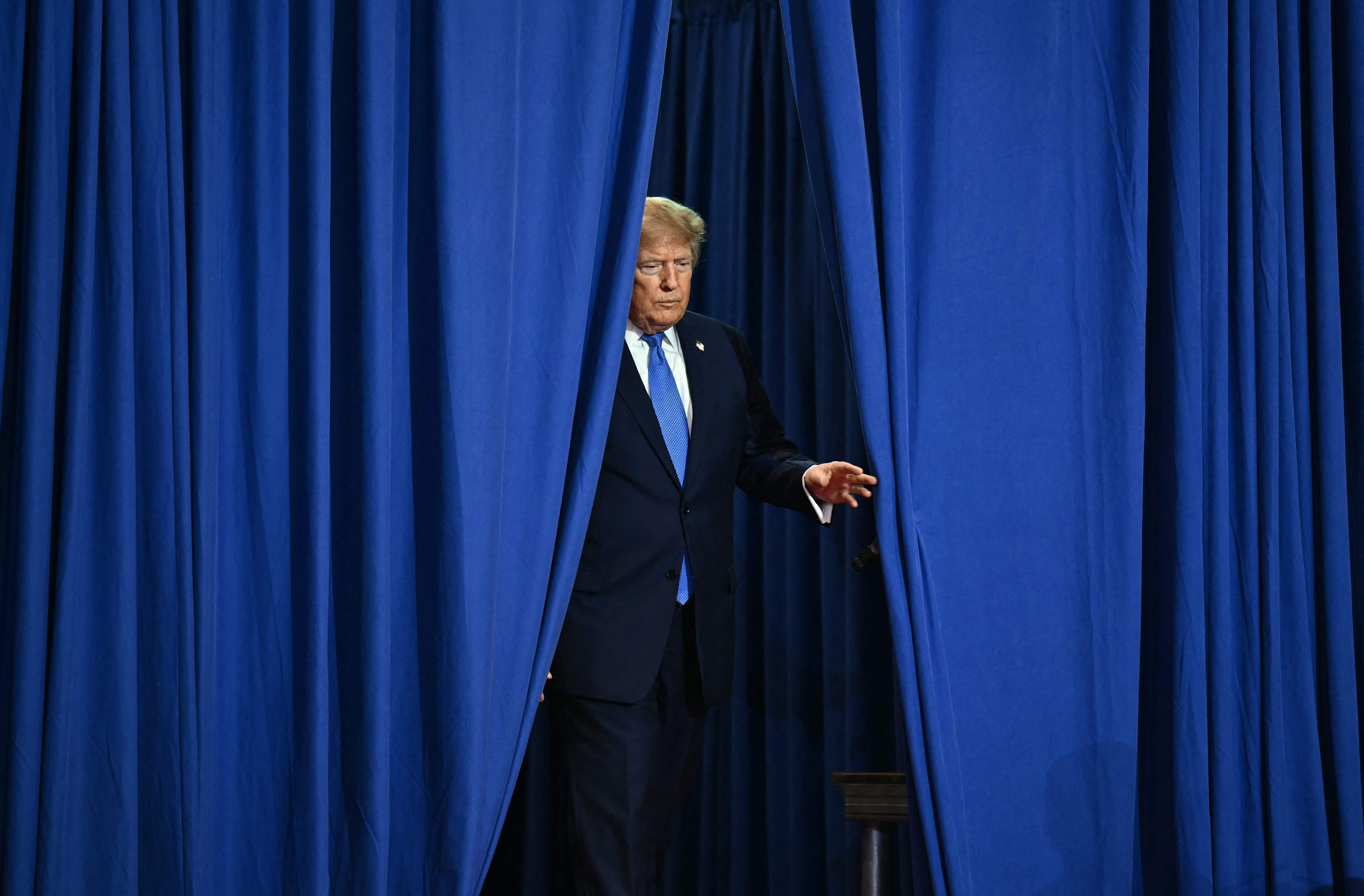 Ein Mann im Anzug tritt durch einen blauen Vorhang auf eine Bühne.