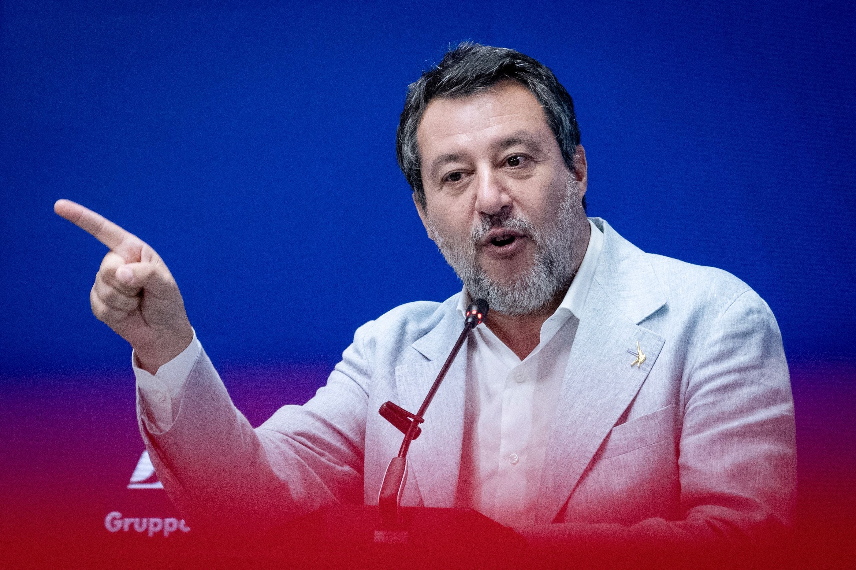 Ein Mann im hellen Anzug spricht gestikulierend am Mikrofon vor blauem Hintergrund.