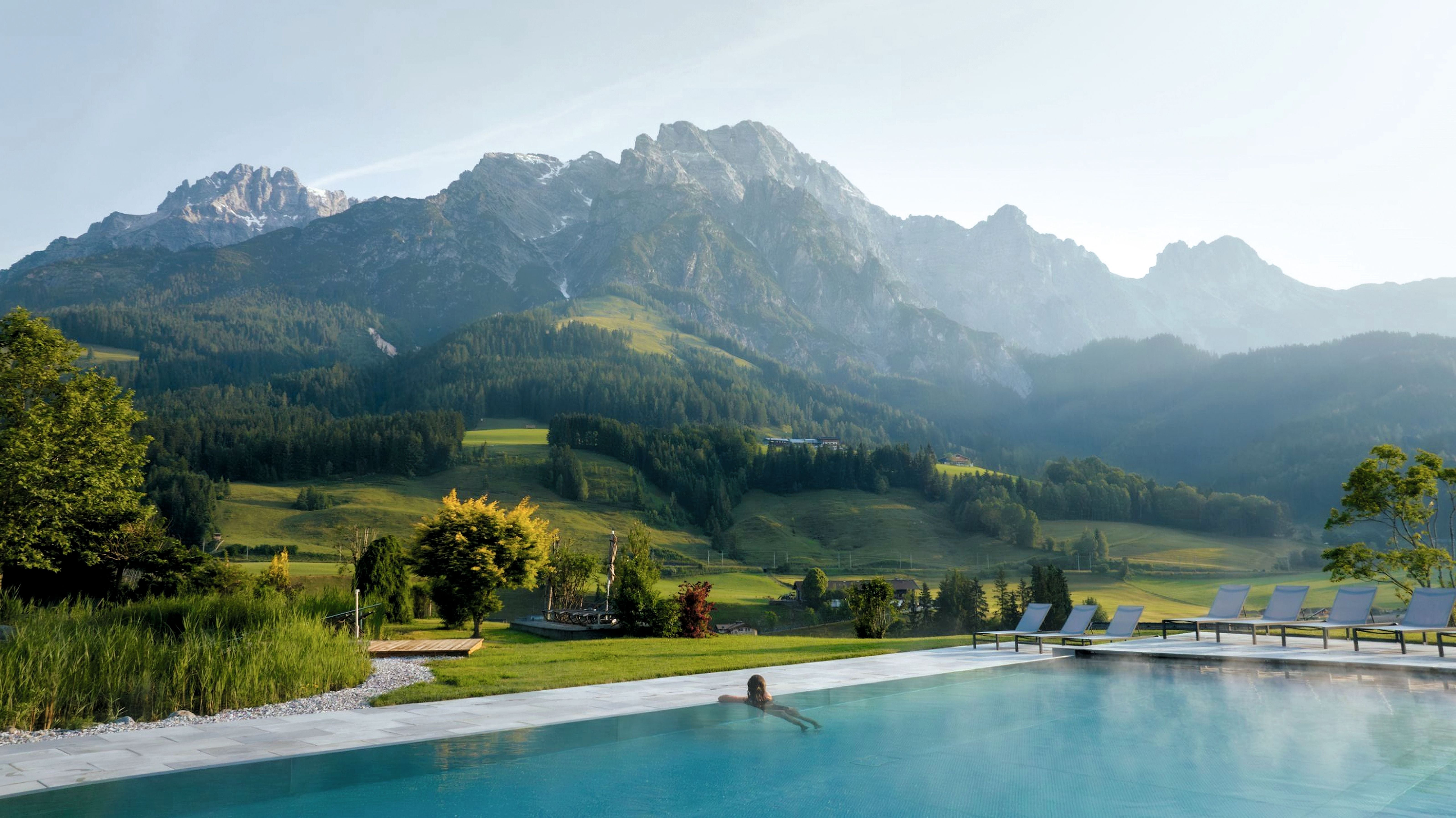 Eine Person schwimmt in einem Pool mit Blick auf grüne Hügel und beeindruckende Berge.
