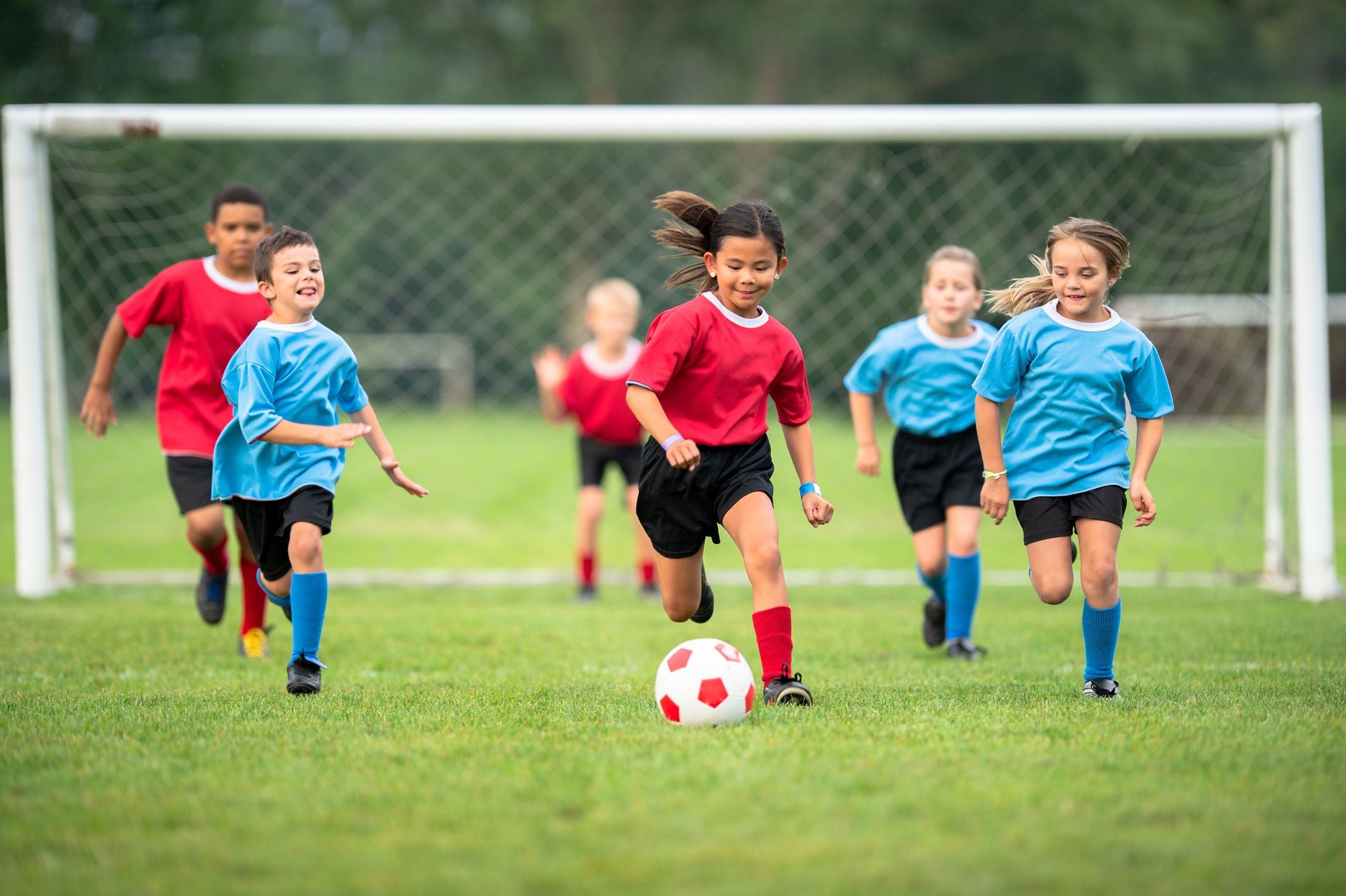 Kinder spielen Fußball, ein Mädchen im roten Trikot läuft mit dem Ball vor mehreren Kindern im blauen Trikot.