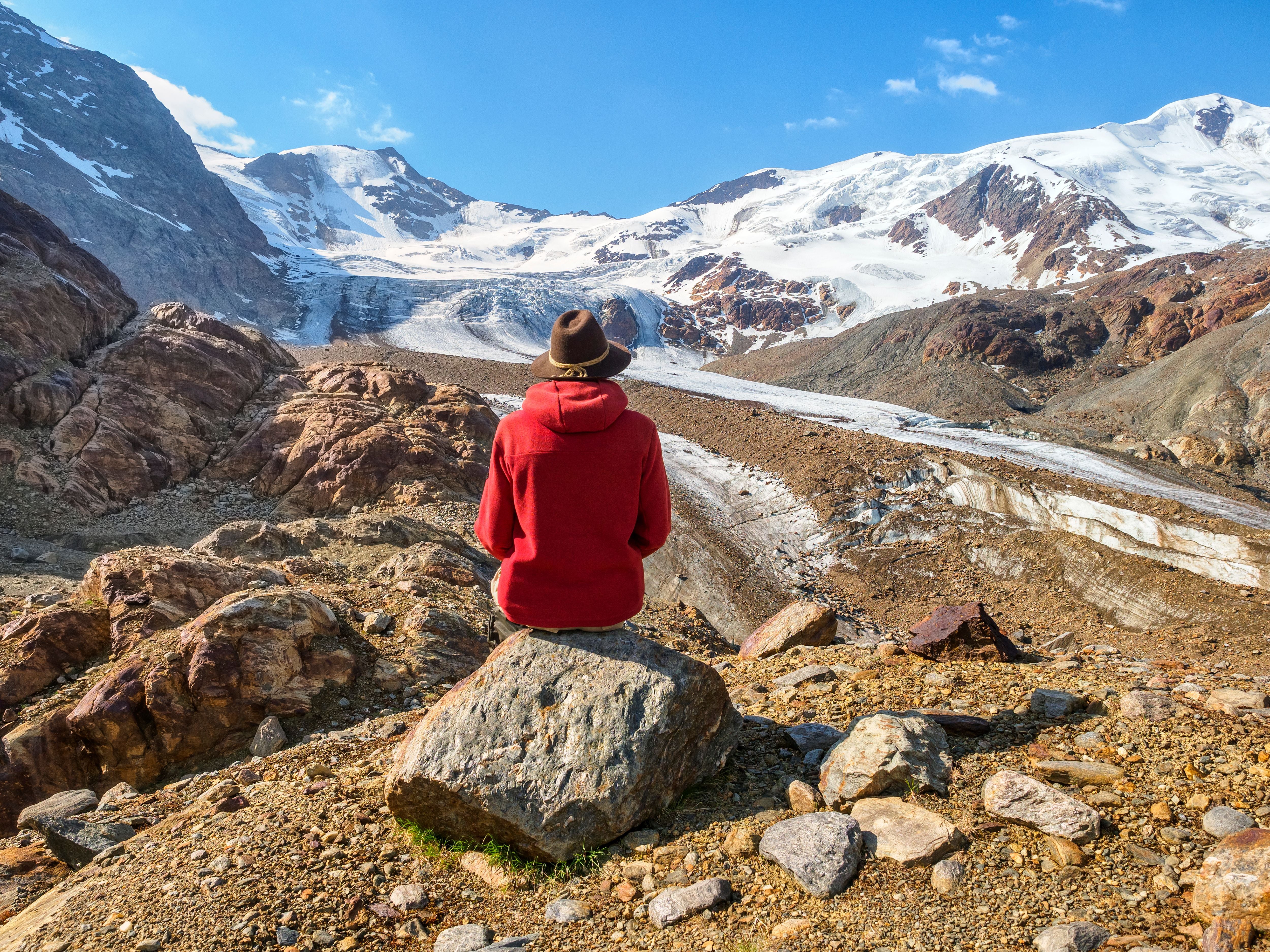 Person mit rotem Hoodie sitzt auf Felsen und blickt auf schneebedeckte Berge und Gletscher.