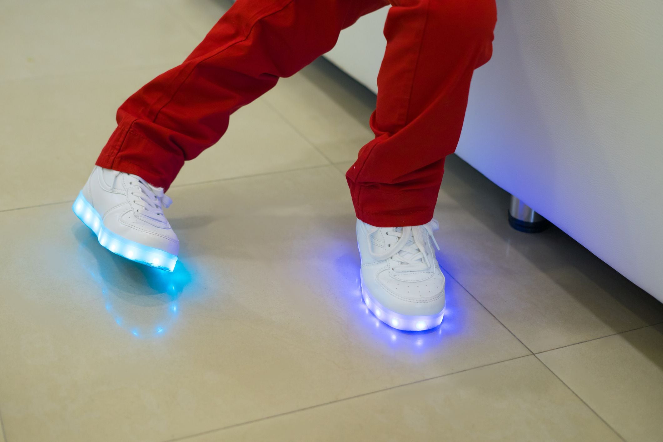 Kind trägt rote Hose und weiße LED-Sneaker mit leuchtenden Sohlen auf Fliesenboden.