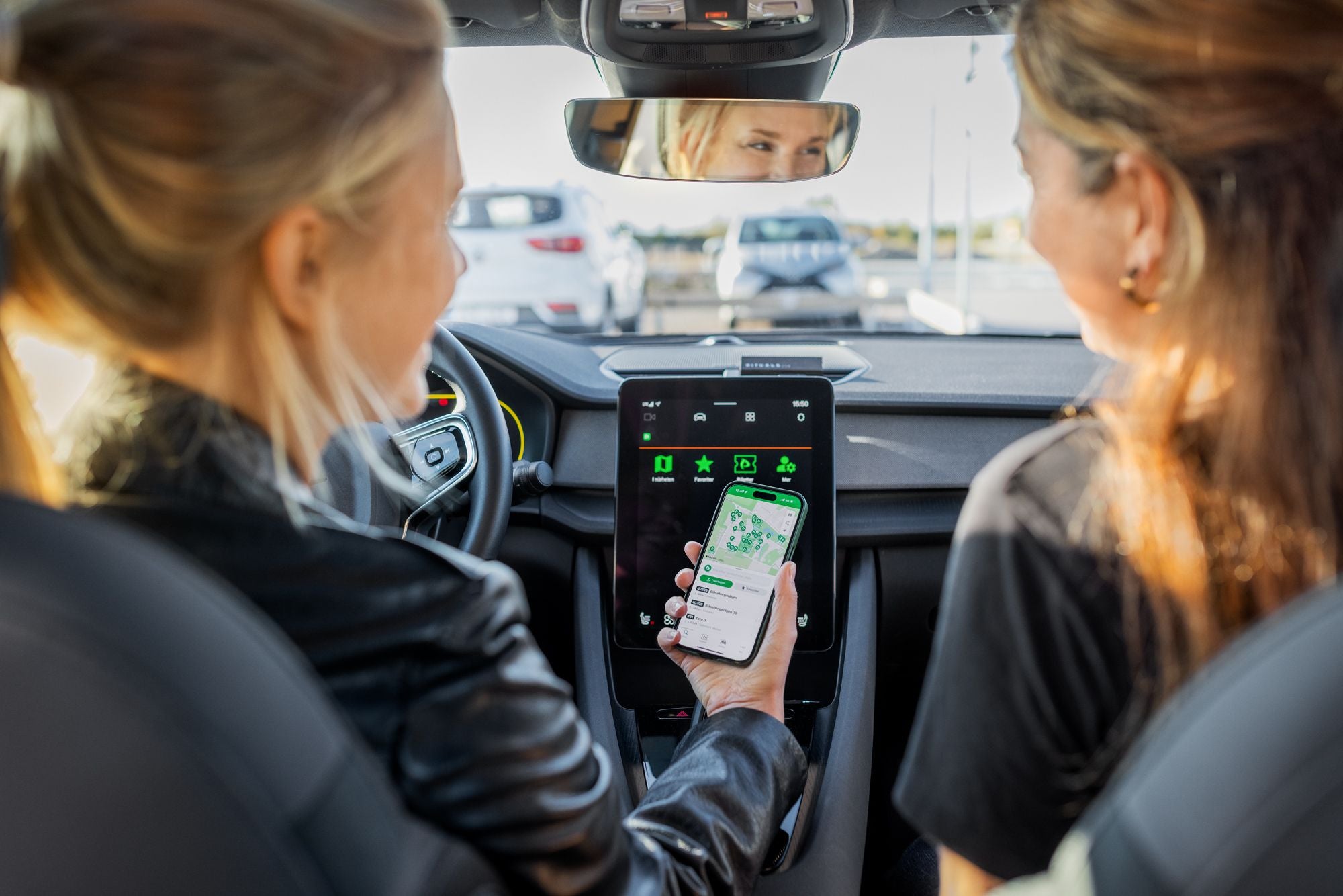 Zwei Frauen sitzen im Auto, eine hält ihr Smartphone mit geöffneter Parkplatz-App.