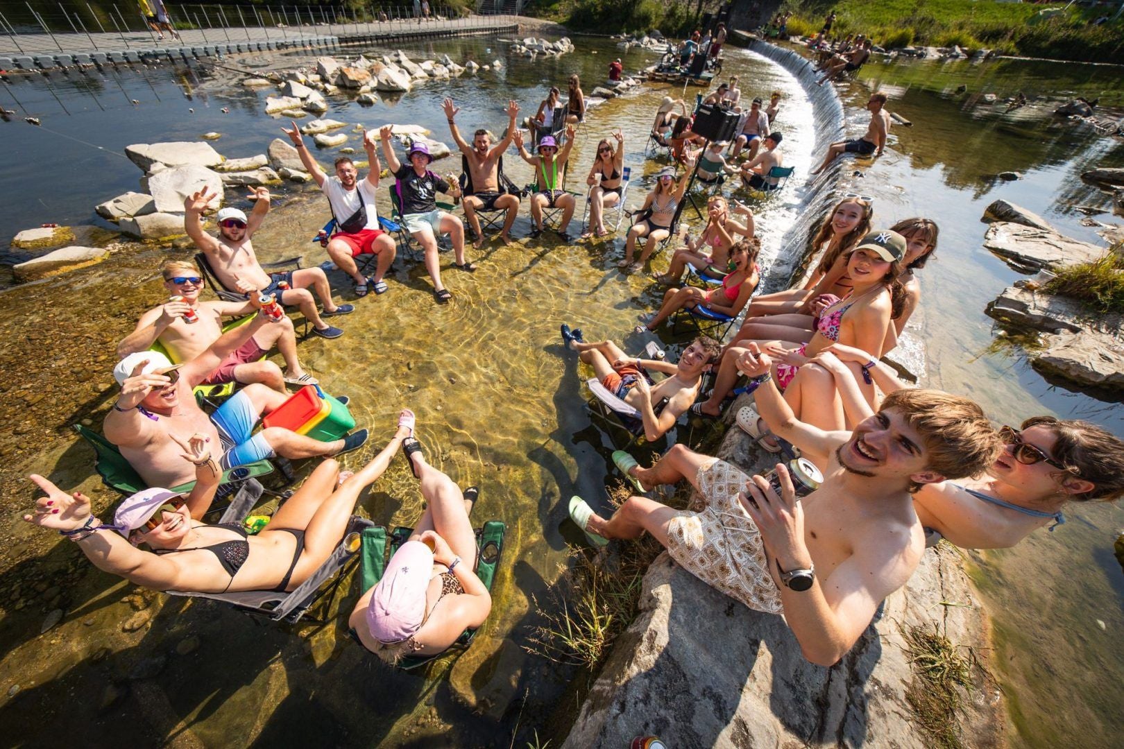 Junge Leute feiern fröhlich im Fluss auf Campingstühlen bei sonnigem Wetter.