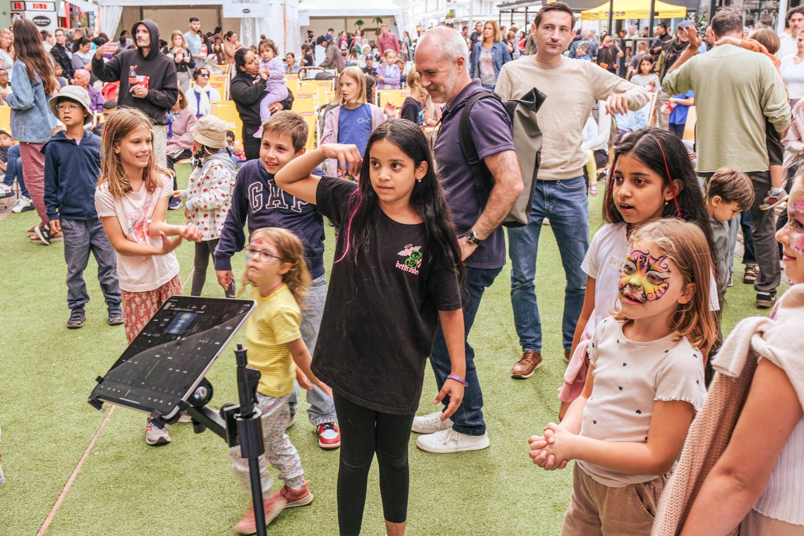 Mehrere Kinder spielen und tanzen fröhlich auf einem Landstraßenfest mit vielen Menschen im Hintergrund.