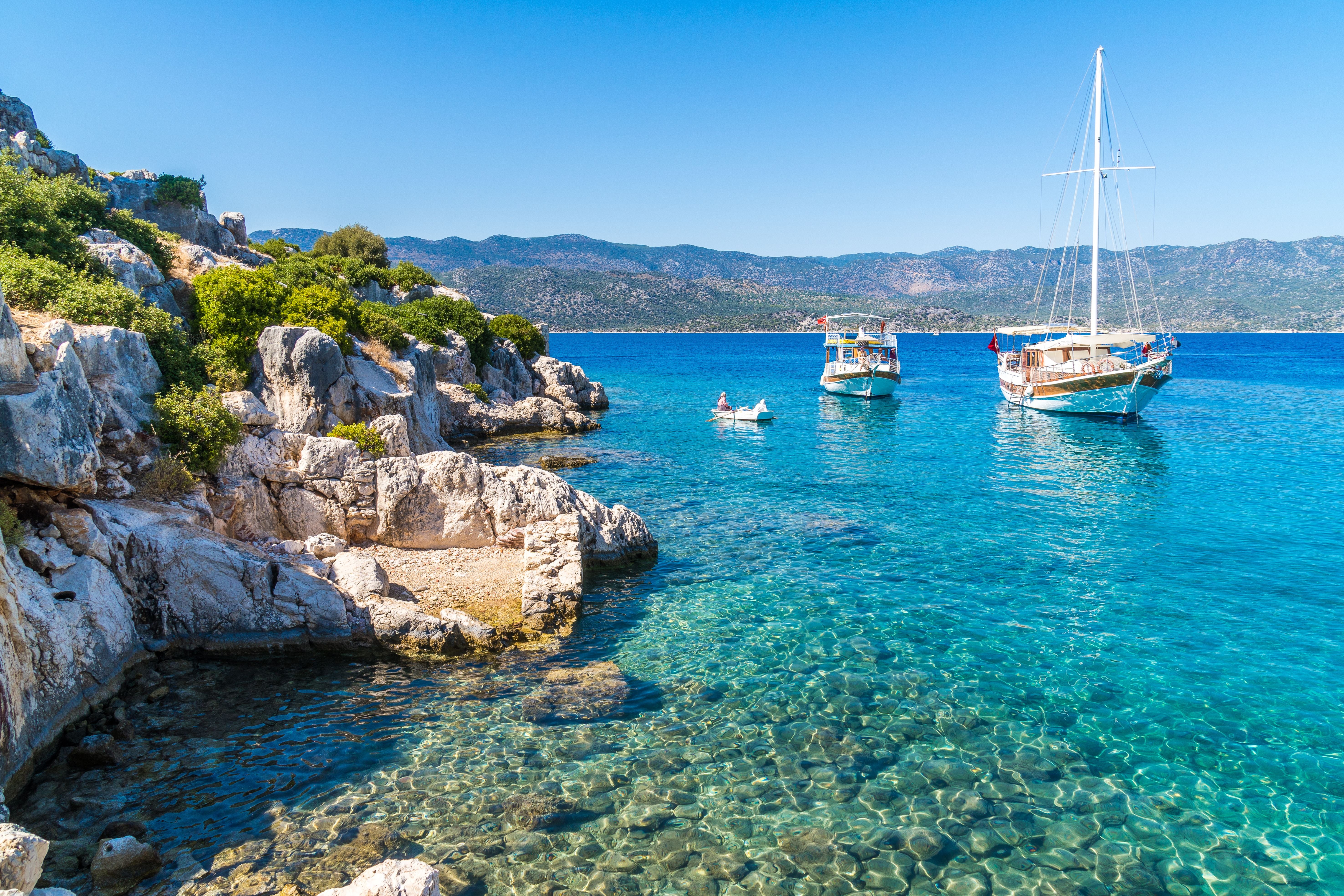 Klares blaues Meer mit Felsen, zwei Booten und einem kleinen Ruderboot vor einer Küste.