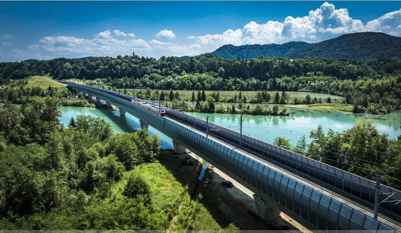 Ein Zug fährt über eine moderne Brücke, umgeben von Fluss, Wald und Bergen bei klarem Himmel.