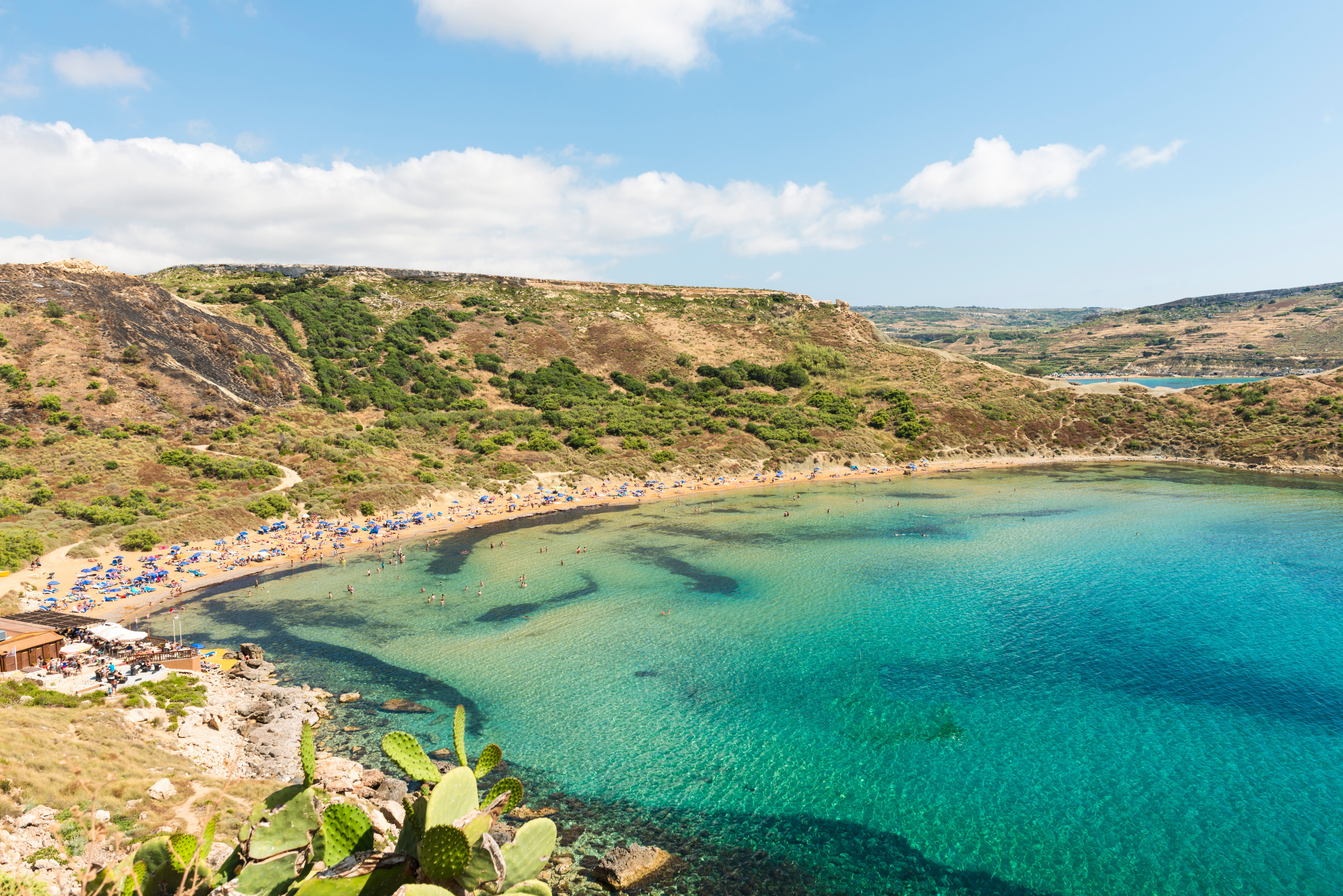 Blick auf den Golden Bay Strand auf Malta mit vielen Badegästen und blauen Sonnenschirmen.