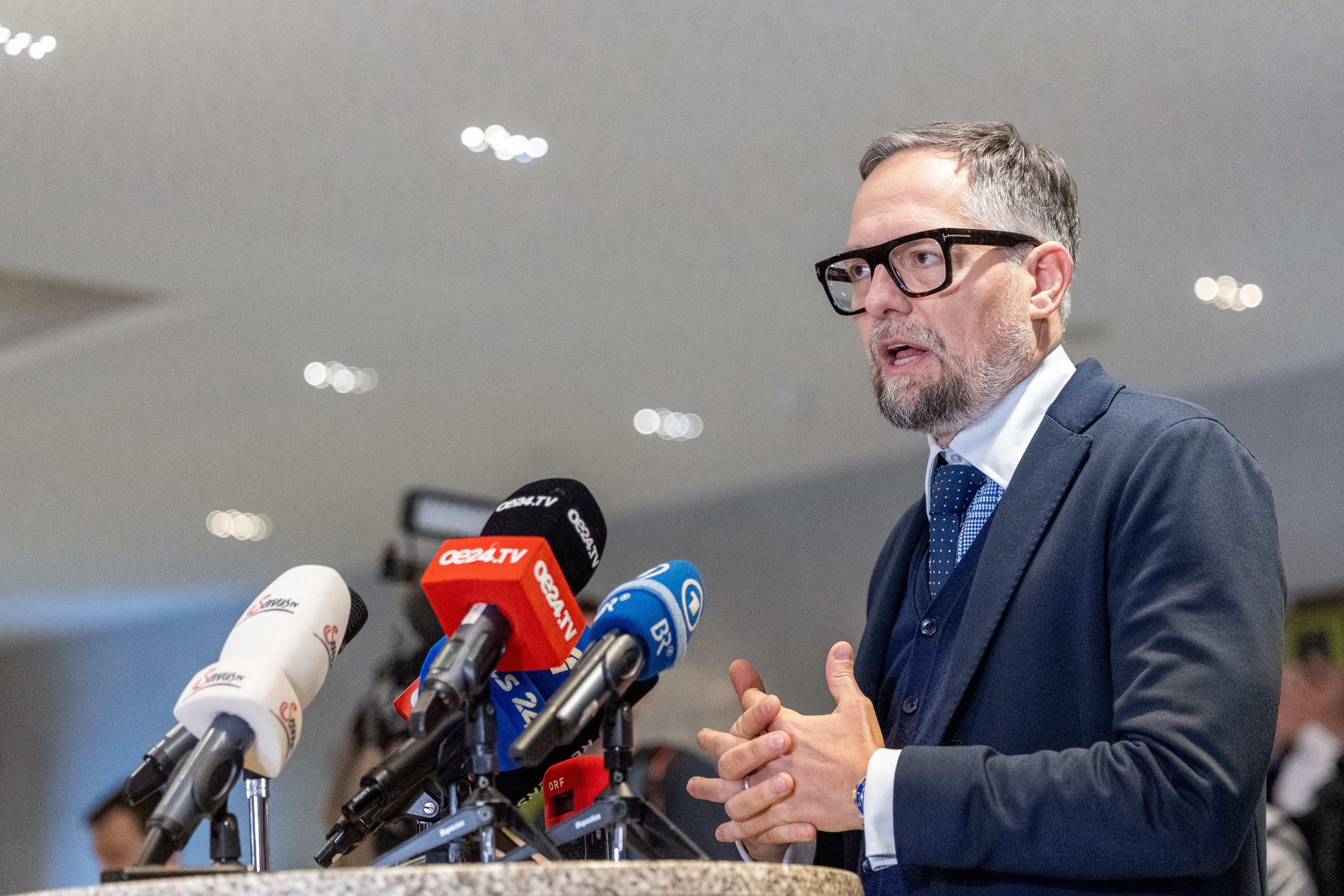 Ein Mann im Anzug spricht bei einer Pressekonferenz vor mehreren Mikrofonen.