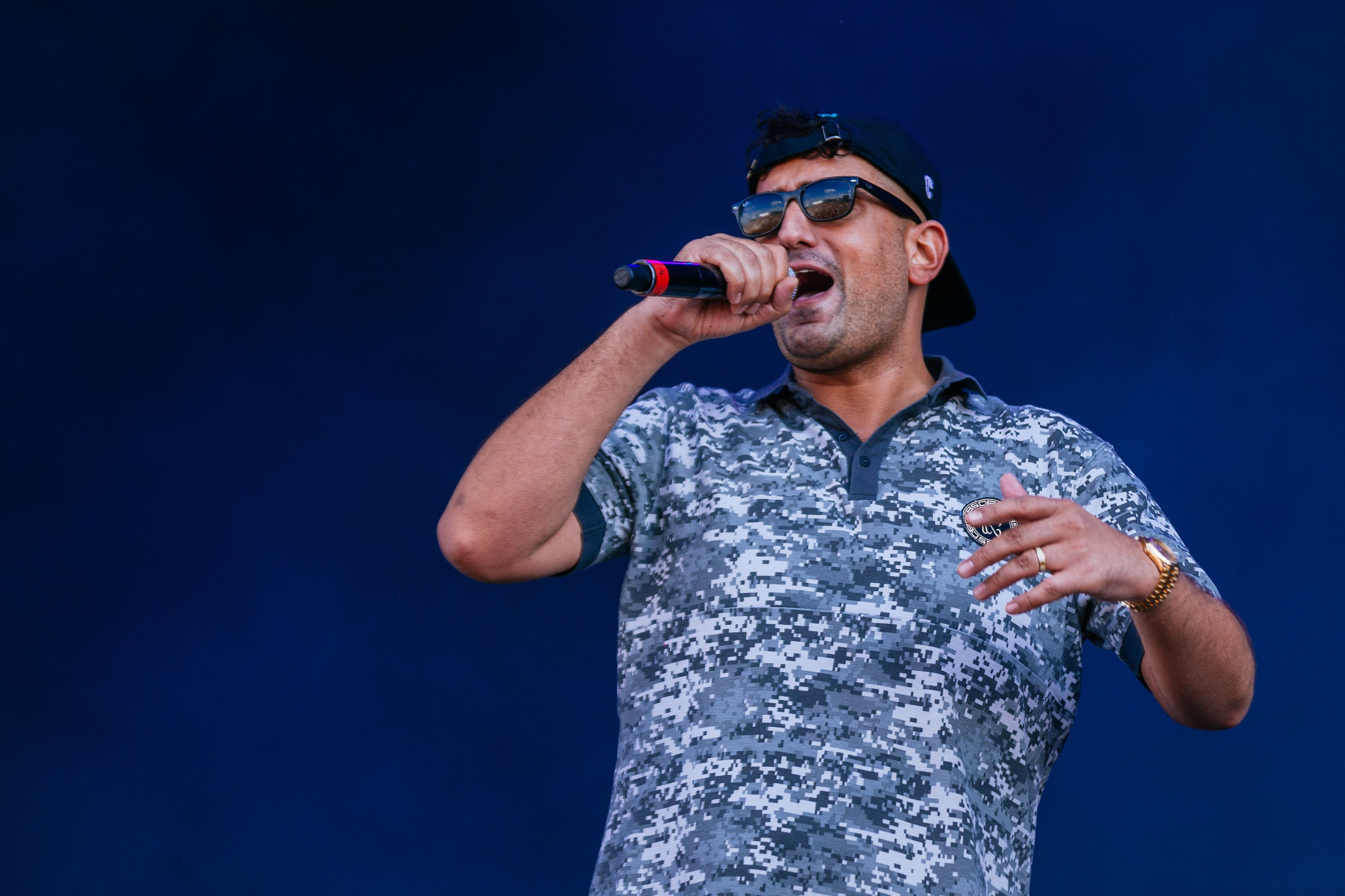 Ein Rapper mit Sonnenbrille und Basecap rappt auf der Bühne vor blauem Hintergrund.