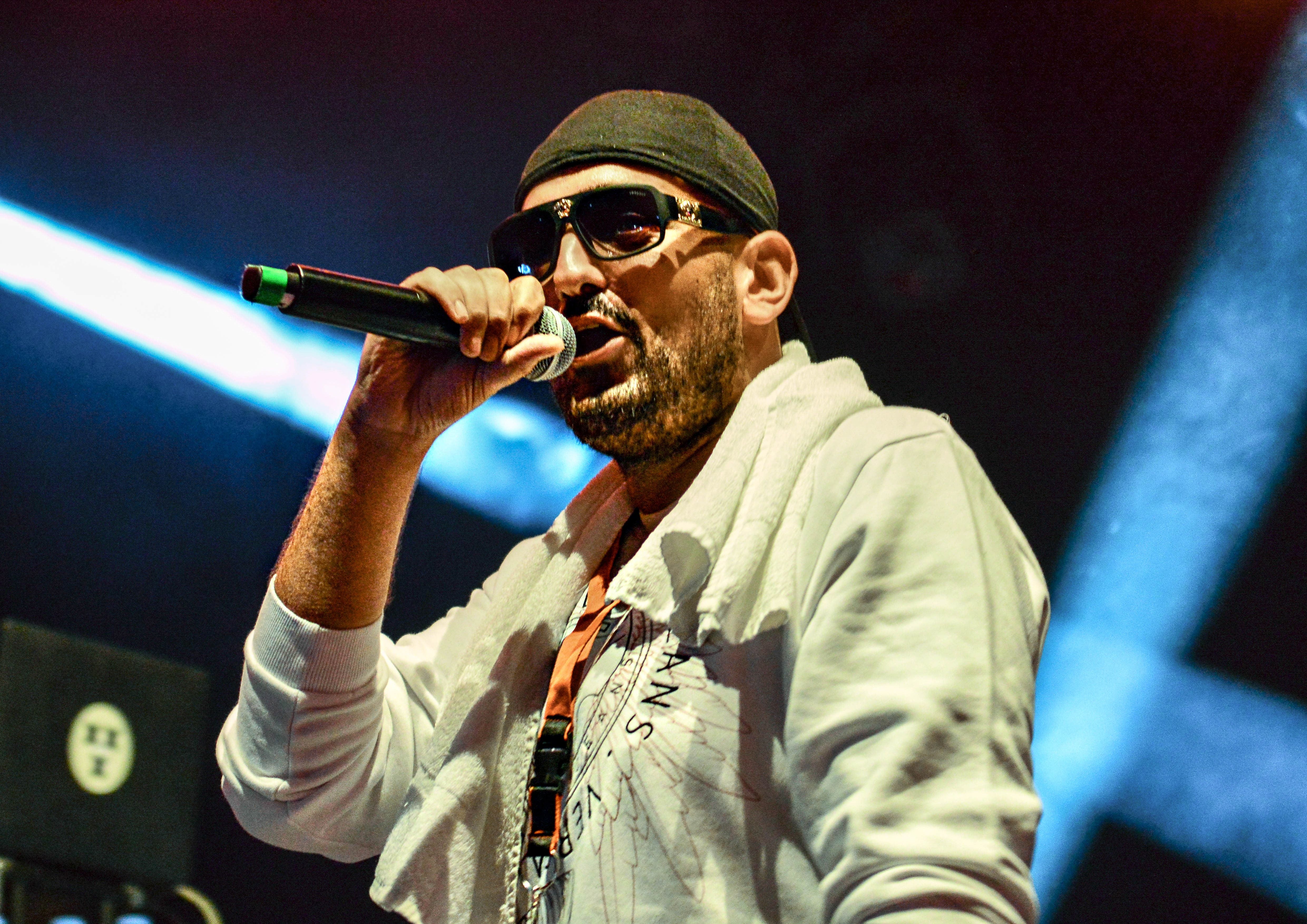 Ein Rapper mit Sonnenbrille und Mütze performt mit Mikrofon auf einer Bühne.
