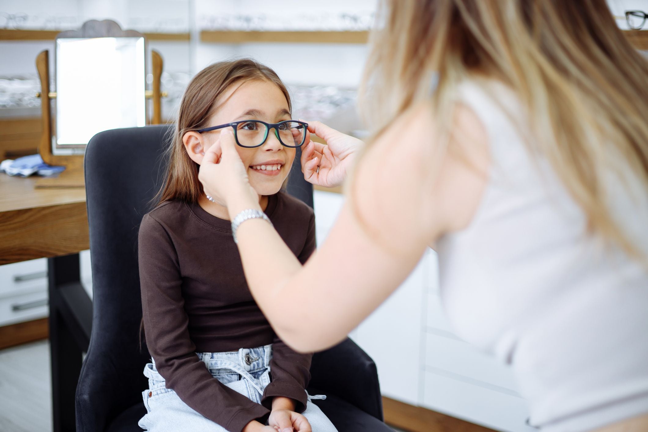 Eine Frau setzt einem lächelnden Mädchen eine Brille im Optikerladen auf.