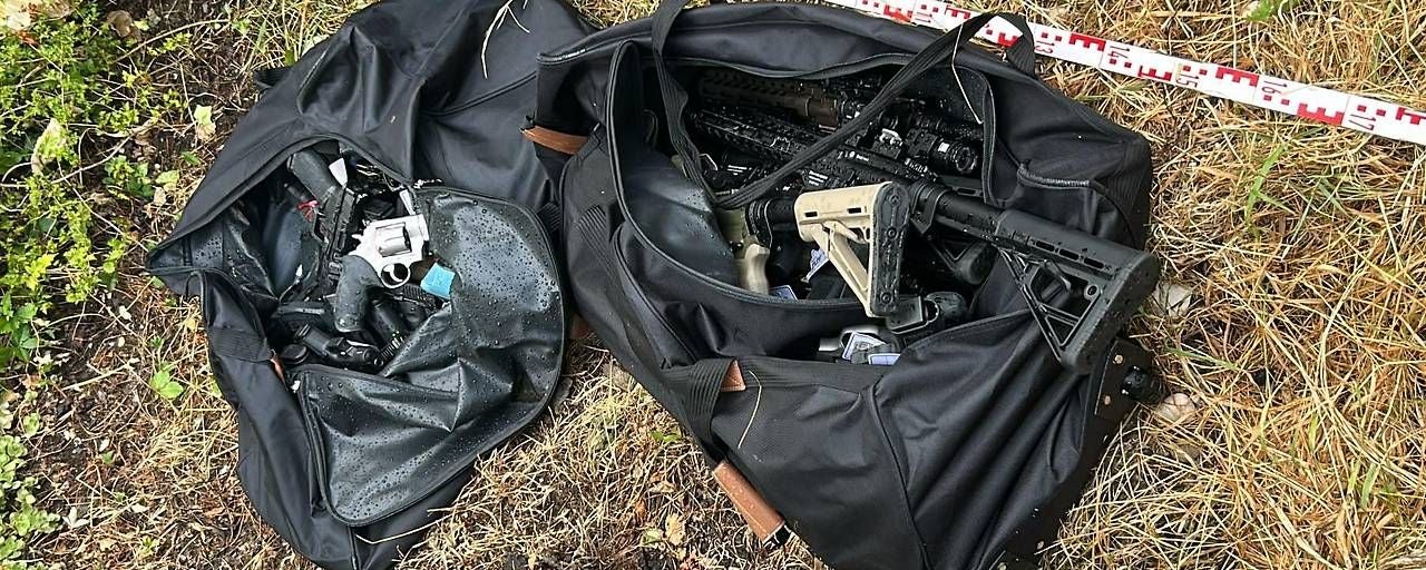 Zwei offene Taschen voller Schusswaffen liegen auf trockenem Gras neben einem Maßband.