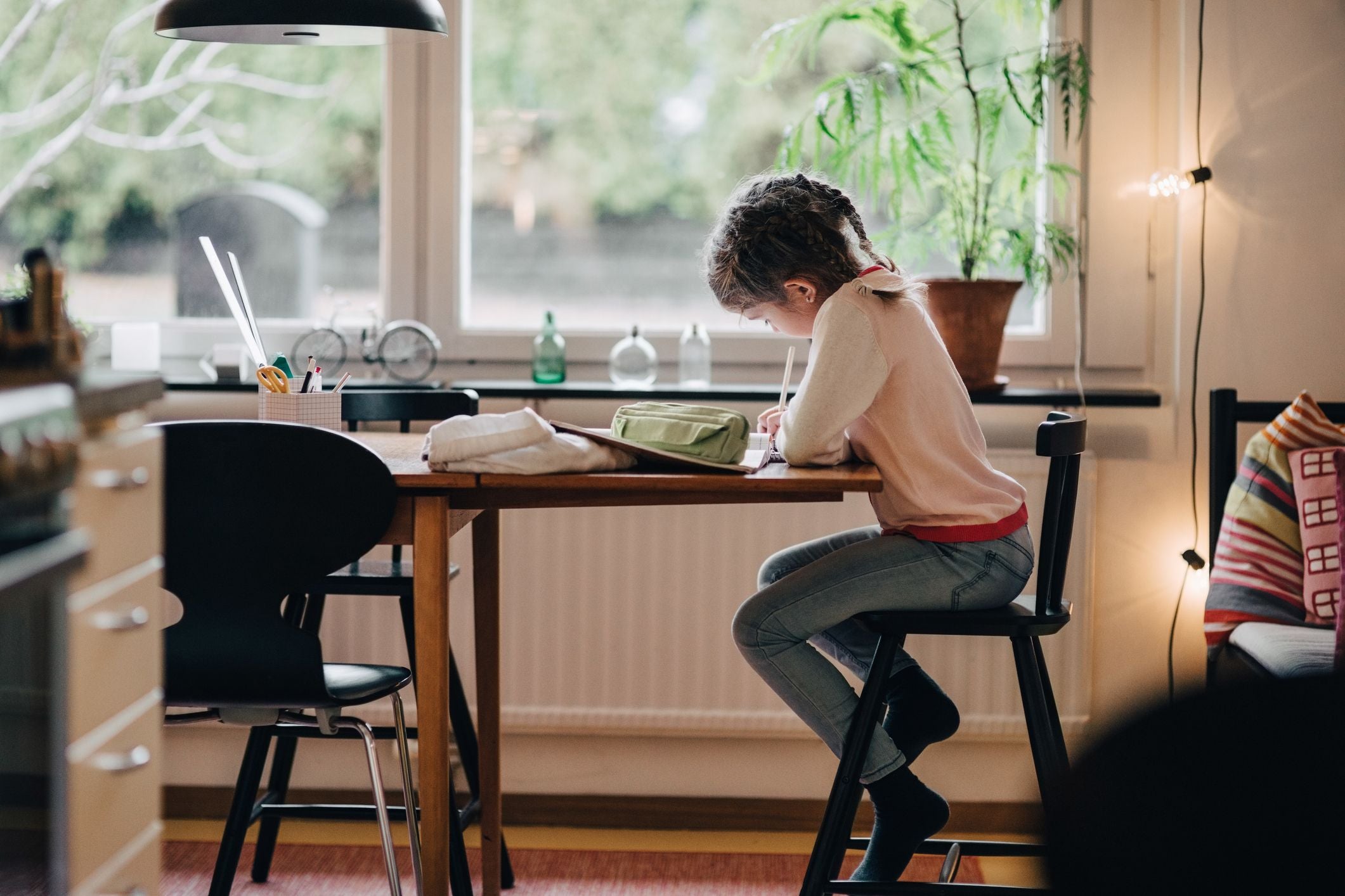 Kind sitzt am Tisch und schreibt, Schulmaterial liegt auf dem Tisch, helle Wohnung im Hintergrund.