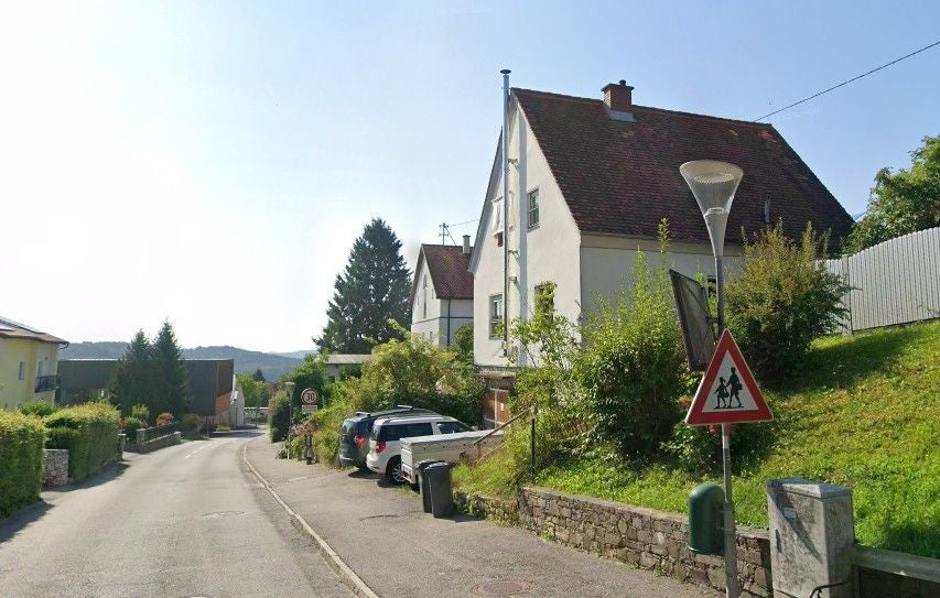 Wohnstraße mit weißem Haus, Autos und einem Verkehrsschild für Kinder am rechten Straßenrand.